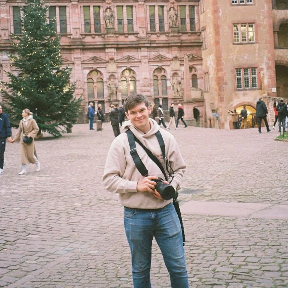 A young man holding a camera, standing in a historic courtyard with a decorated Christmas tree nearby, surrounded by tourists and an ornate stone building with statues and windows.