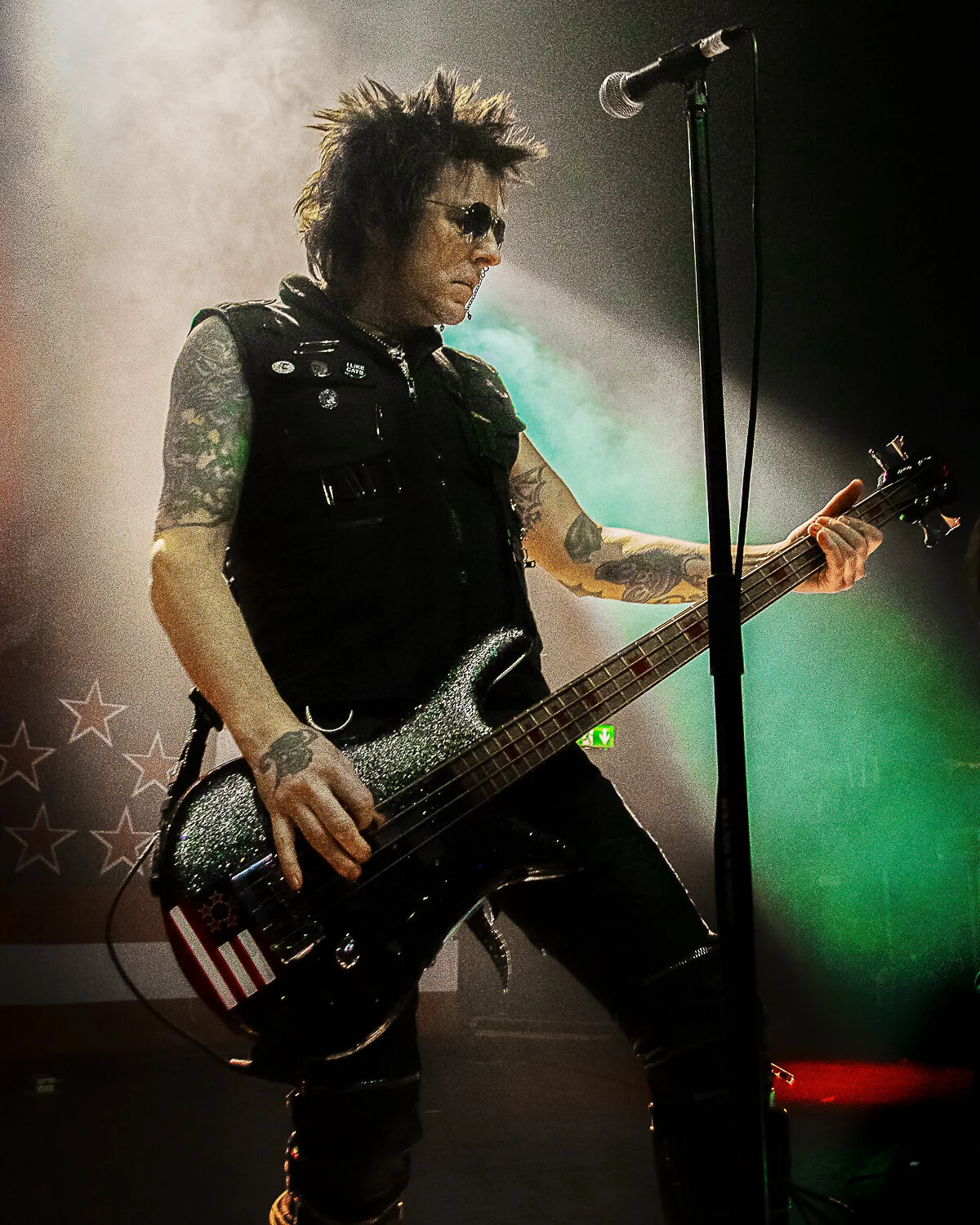 Rachel Bolan playing bass at the microphone, green and white light behind him, Skid Row live in concert at The O2 Forum Kentish Town, January 2019
