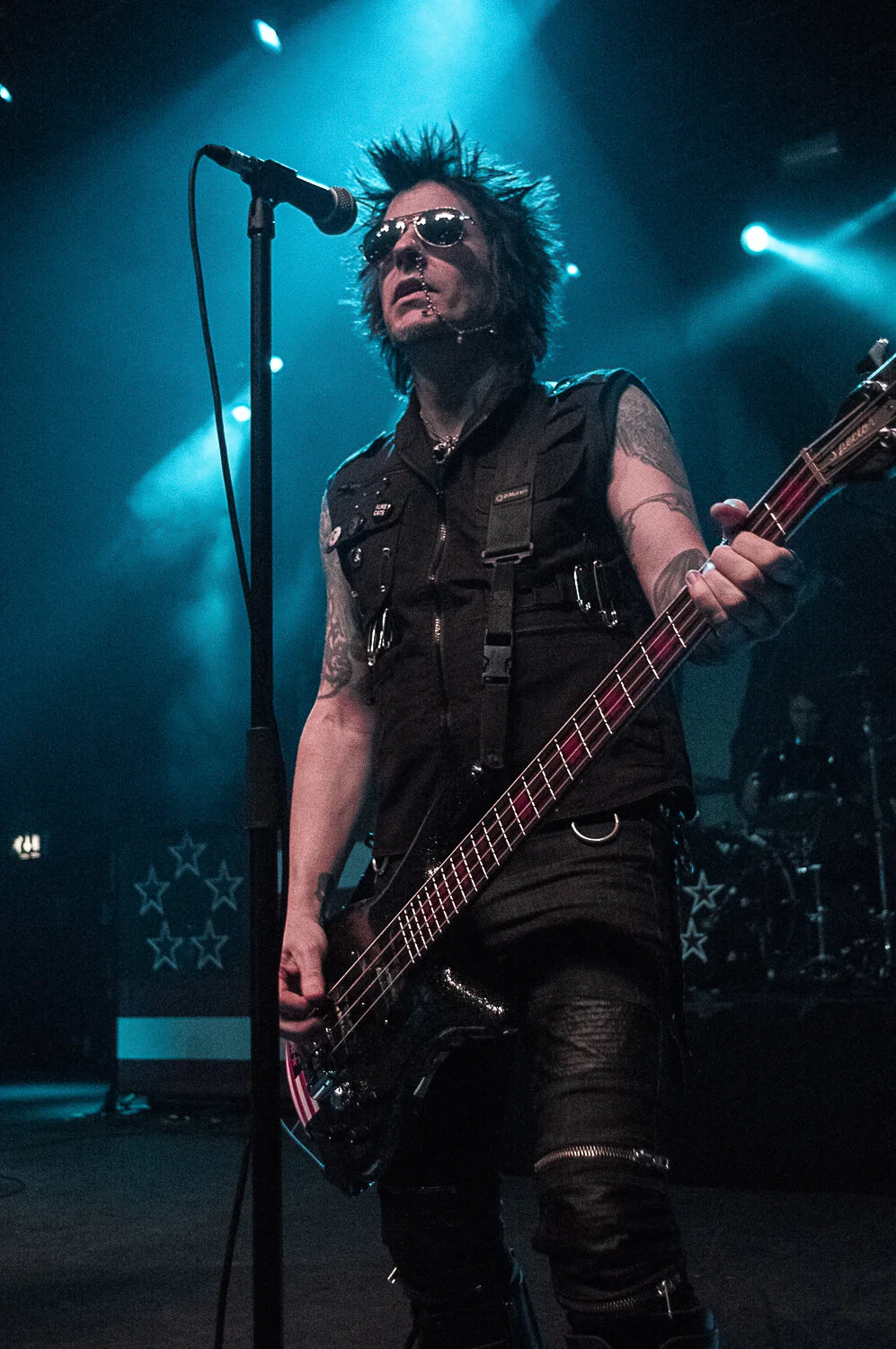 Rachel Bolan playing bass at the microphone under blue stage lights, Skid Row live in concert at The O2 Forum Kentish Town, January 2019
