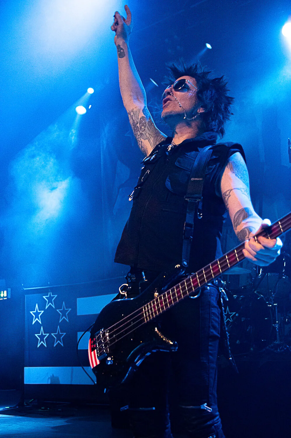 Rachel Bolan raising his arm while playing bass under blue stage lights, Skid Row live in concert at The O2 Forum Kentish Town, January 2019