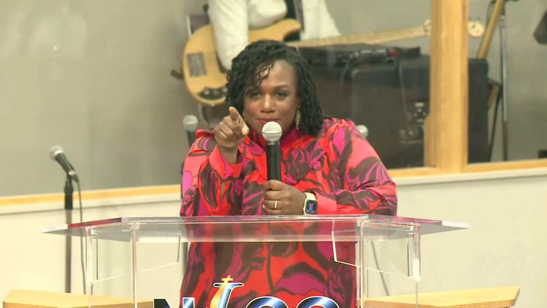 A woman with dark curly hair, wearing a vibrant red and pink patterned dress, is speaking at a church pulpit movie speaker with a microphone in her hand, pointing forward.