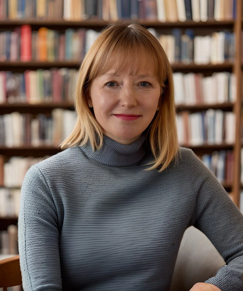 A woman with red hair sitting in front of a bookshelf filled with books, wearing a gray turtleneck sweater.