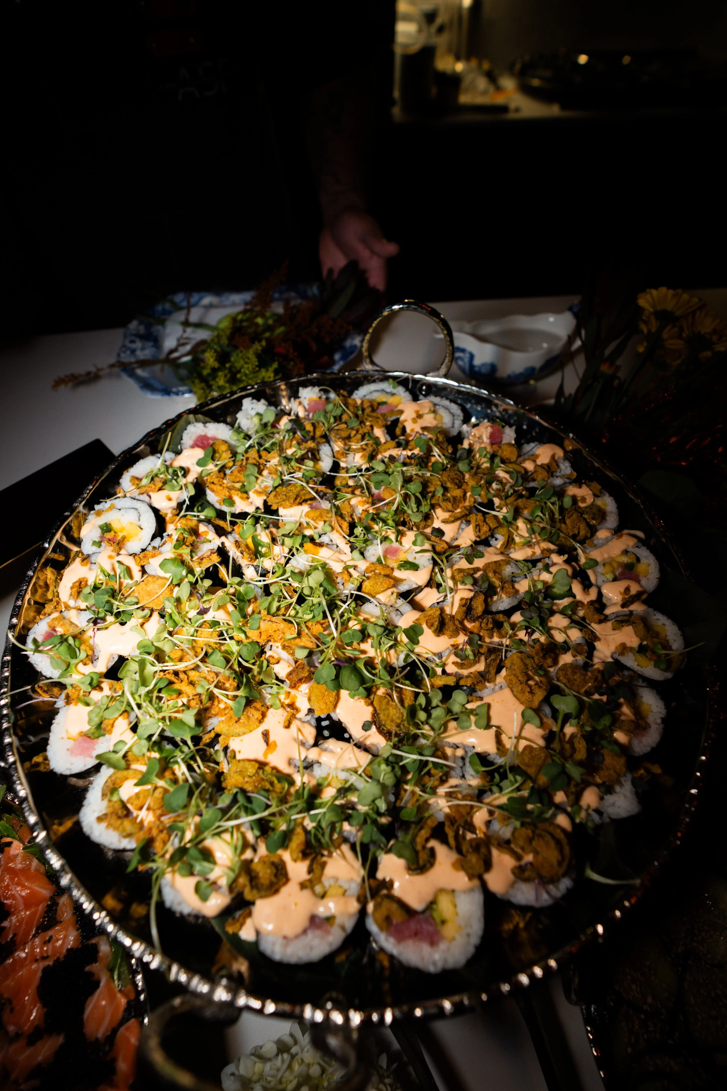 A large silver platter of sushi rolls topped with microgreens and drizzled with orange sauce, displayed at a buffet or catering event.