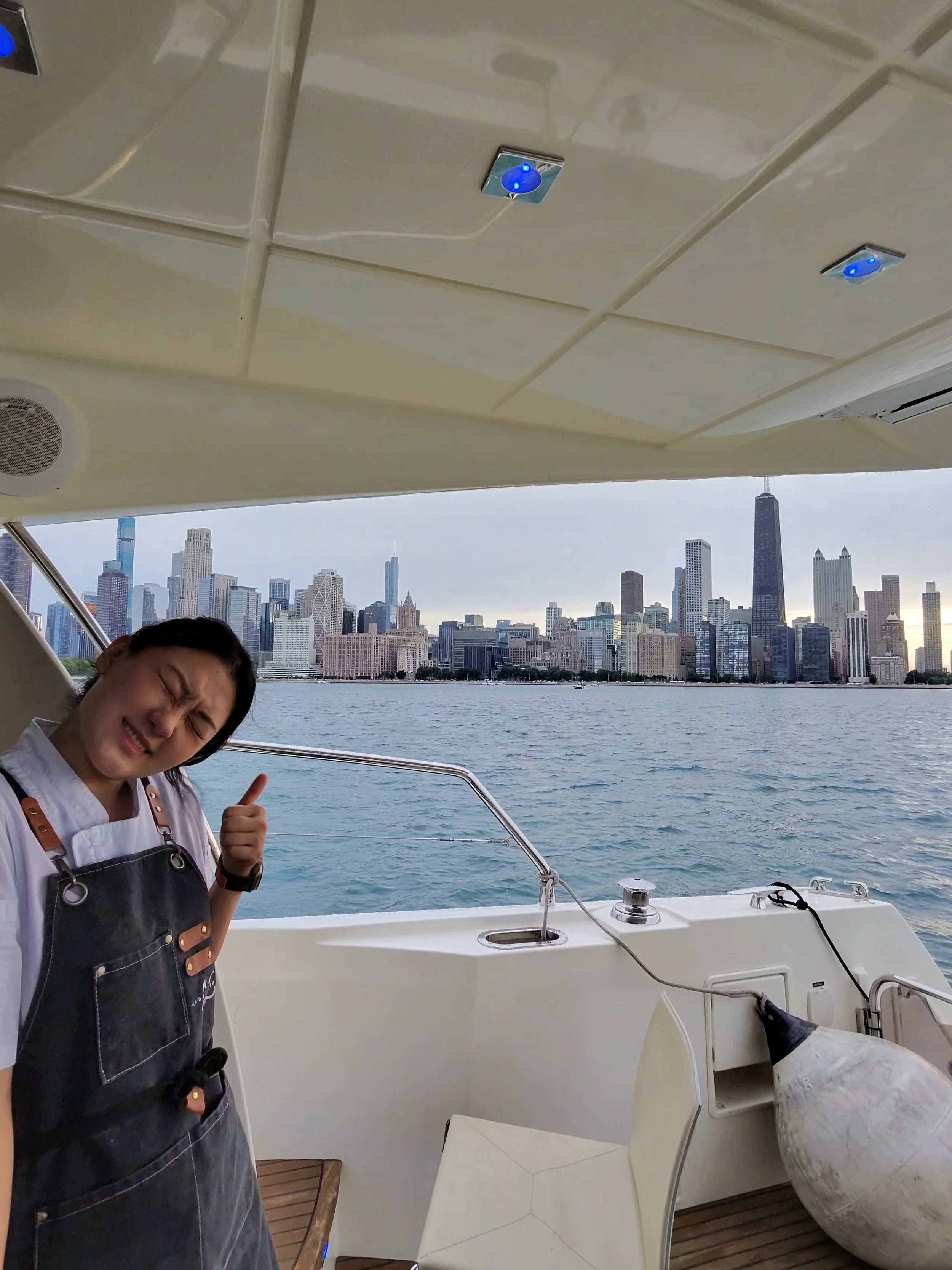Person on a boat with a city skyline in the background, giving a thumbs up and smiling with eyes closed.