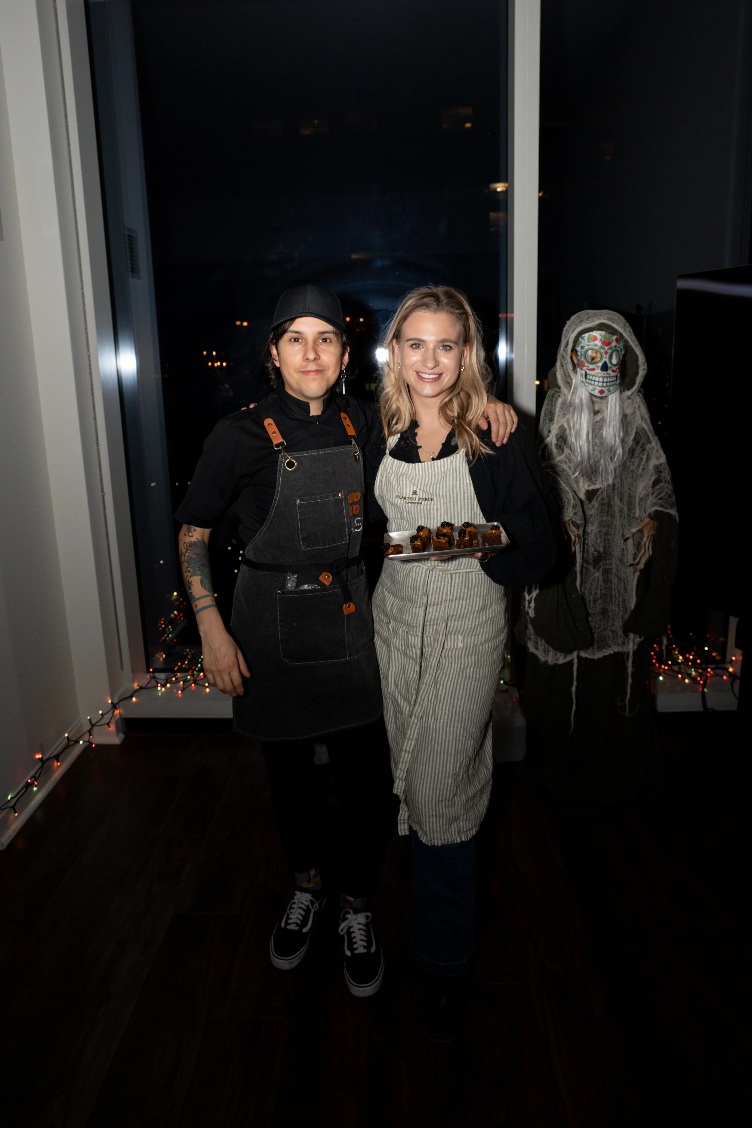 Two people celebrating Halloween, one dressed as a chef and the other as a baker holding a tray of cupcakes, standing next to a spooky skeleton decoration in a decorated indoor setting at night.