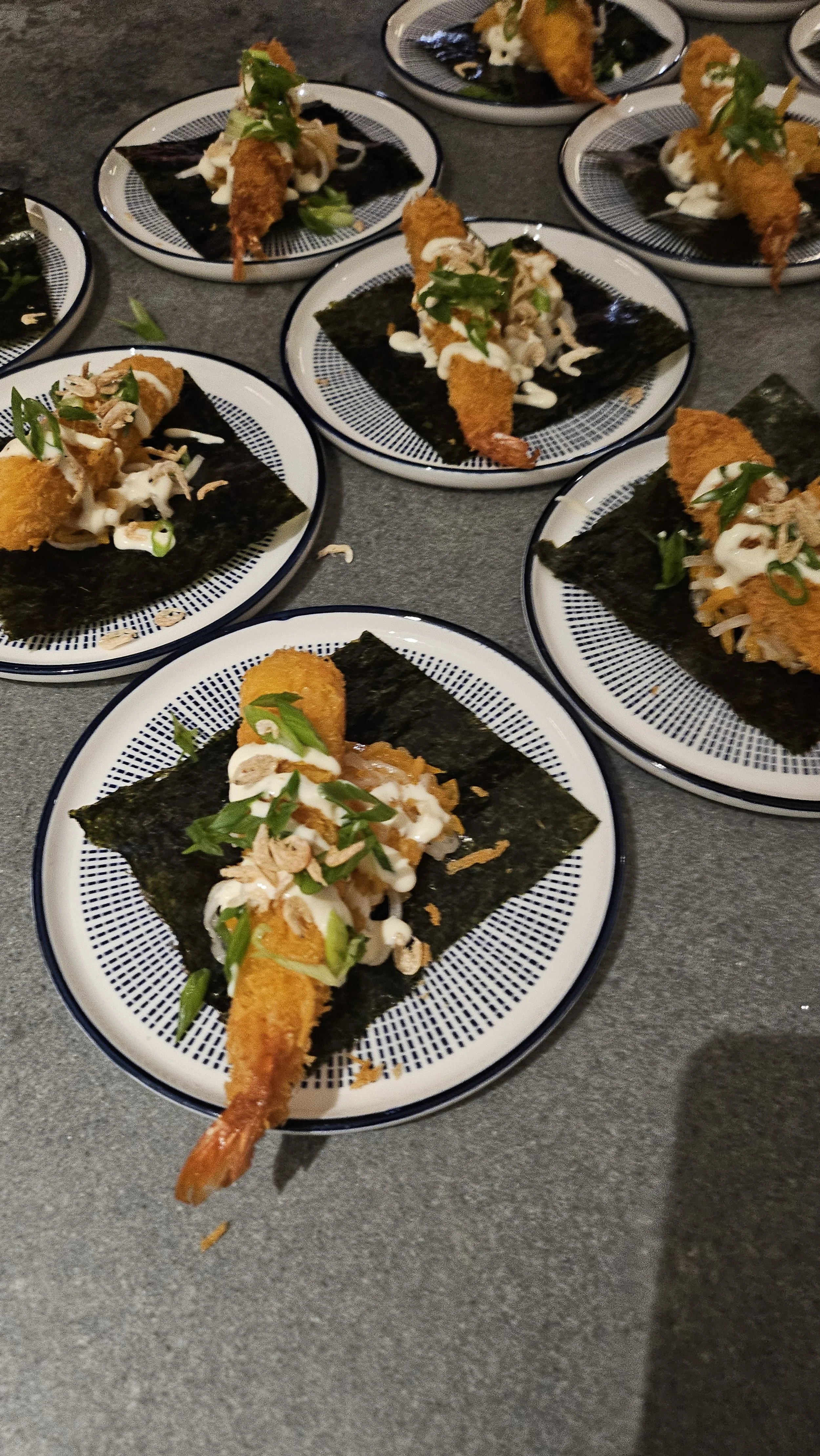 Multiple plates of Japanese tempura shrimp on nori seaweed, garnished with sliced green onions and drizzled with white sauce.