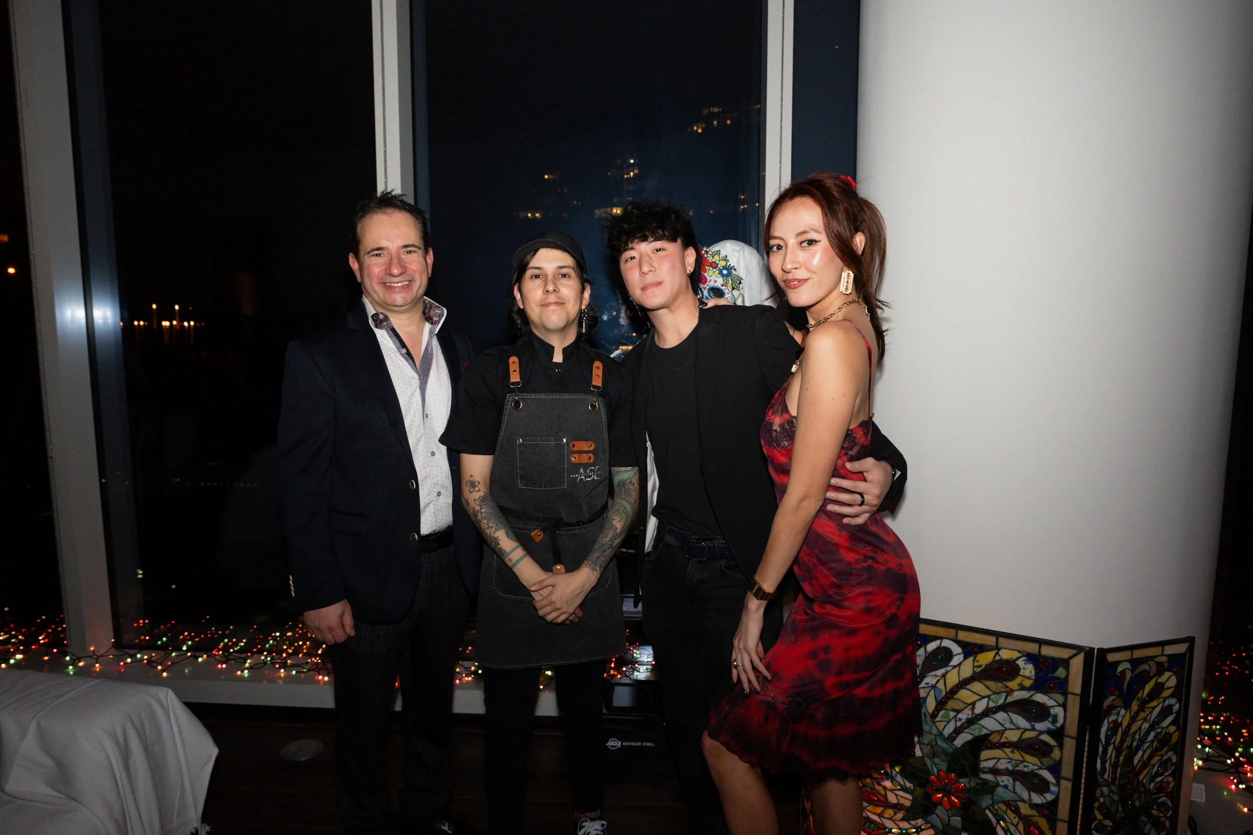 Five people standing together in a high-rise apartment at night, with city lights visible through large windows behind them. They are dressed in semi-formal and casual attire, smiling for the camera.