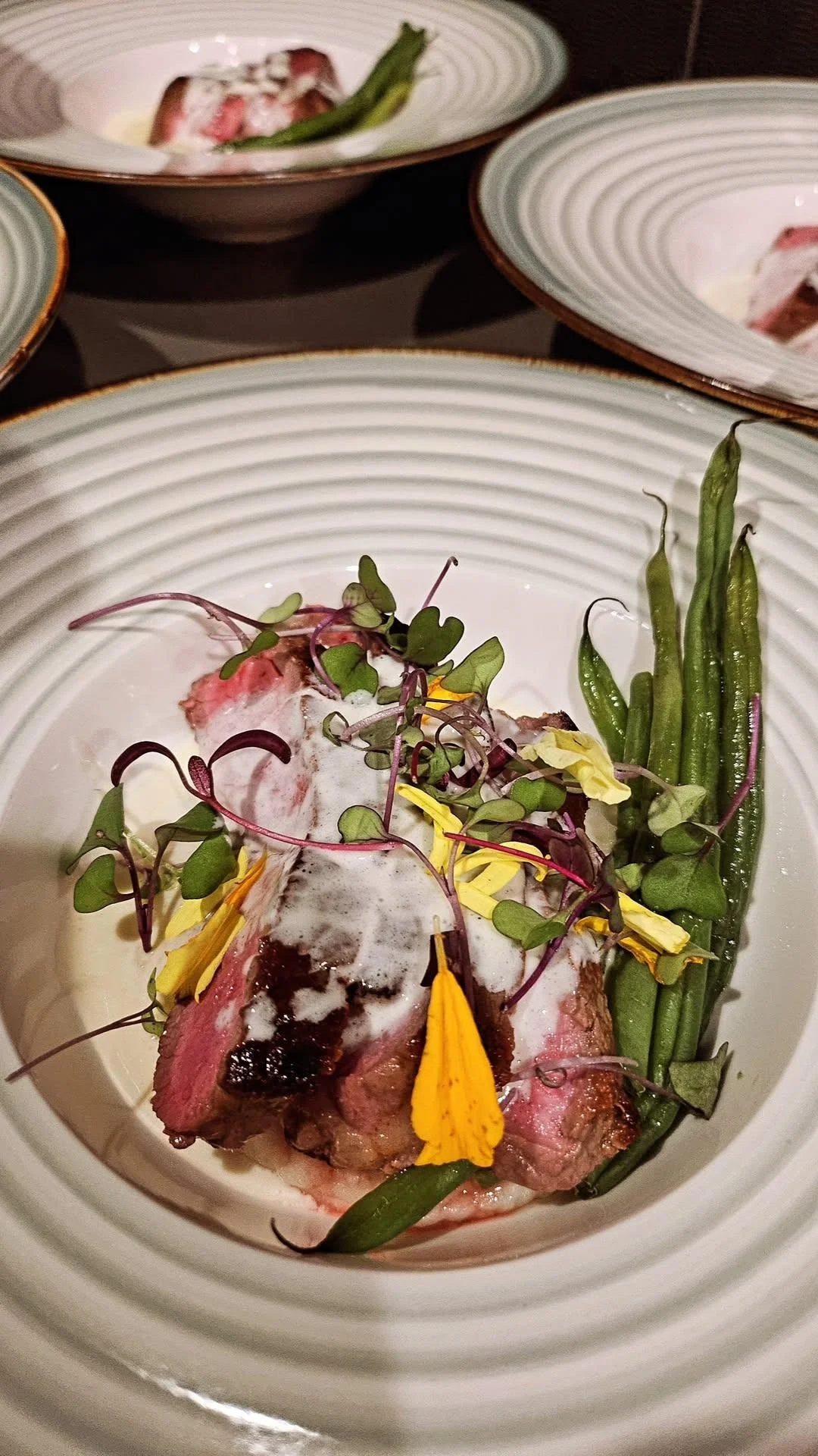 A plated entree featuring cooked meat topped with foamed sauce, microgreens, edible yellow flower petals, green beans, and asparagus on a white plate with green and gold rim.