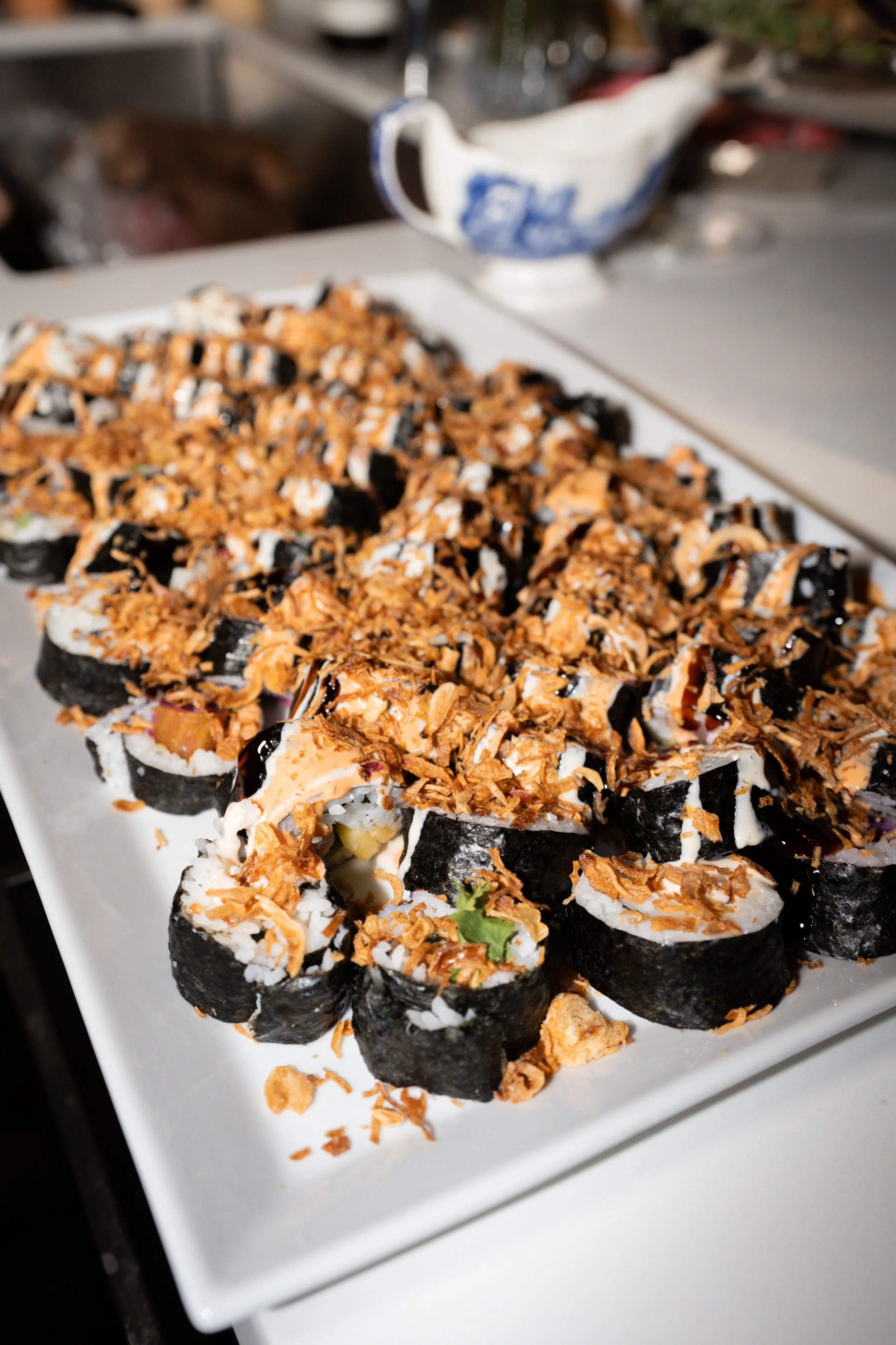 Close-up of a plate of sushi rolls topped with crispy fried onions and drizzled with sauces, on a white rectangular serving dish.