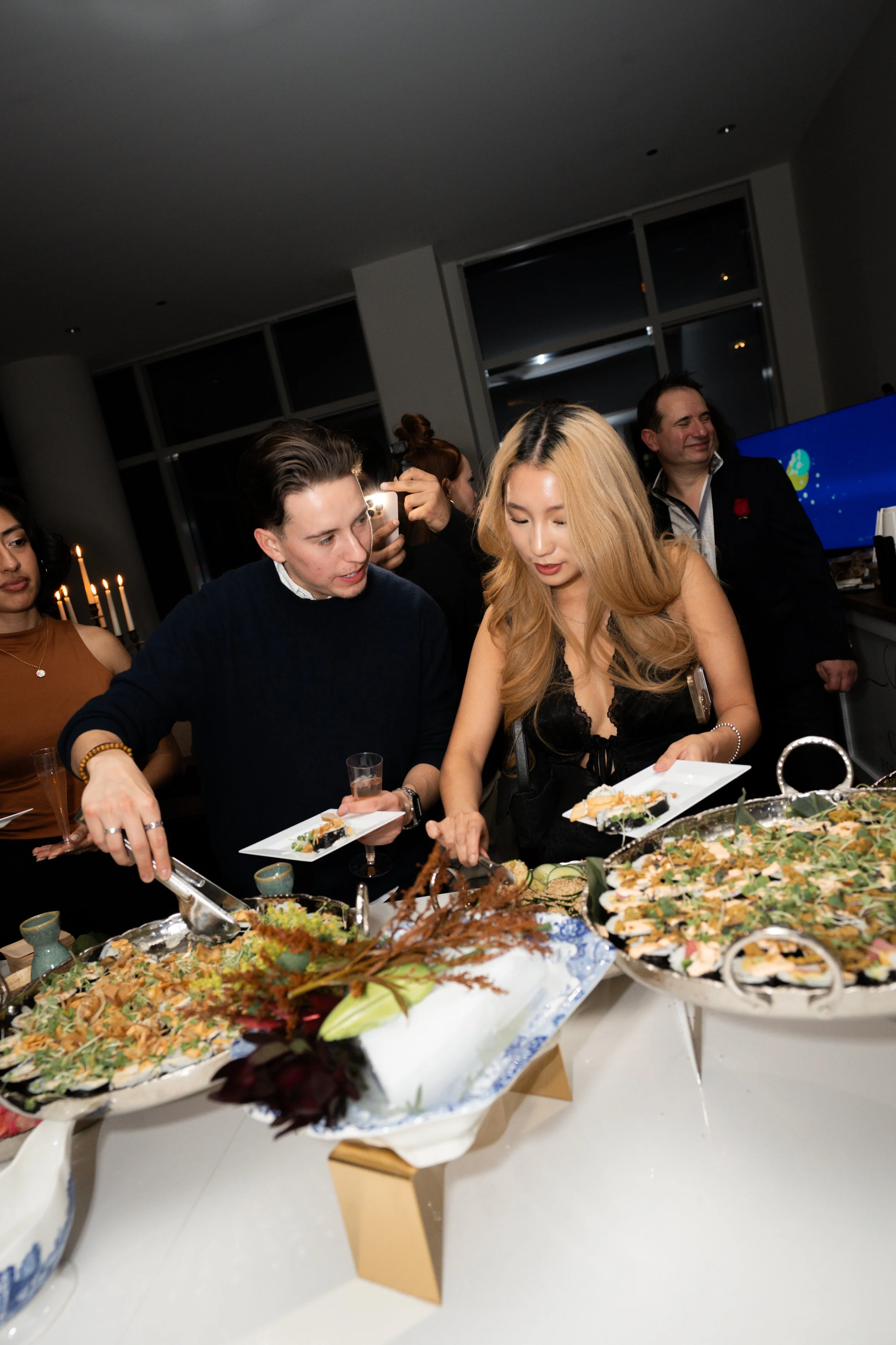 People serving themselves food at a social gathering in a modern indoor setting with large windows.