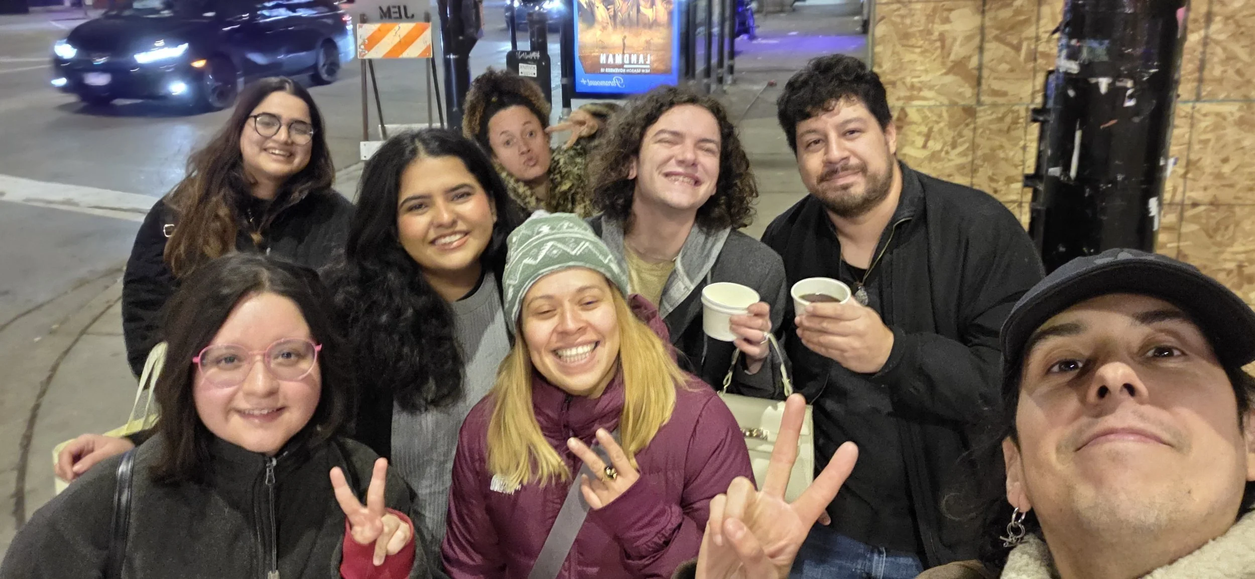 Group of nine friends smiling and making peace signs outside at night, with some holding cups, standing in front of a wooden wall on a city sidewalk.