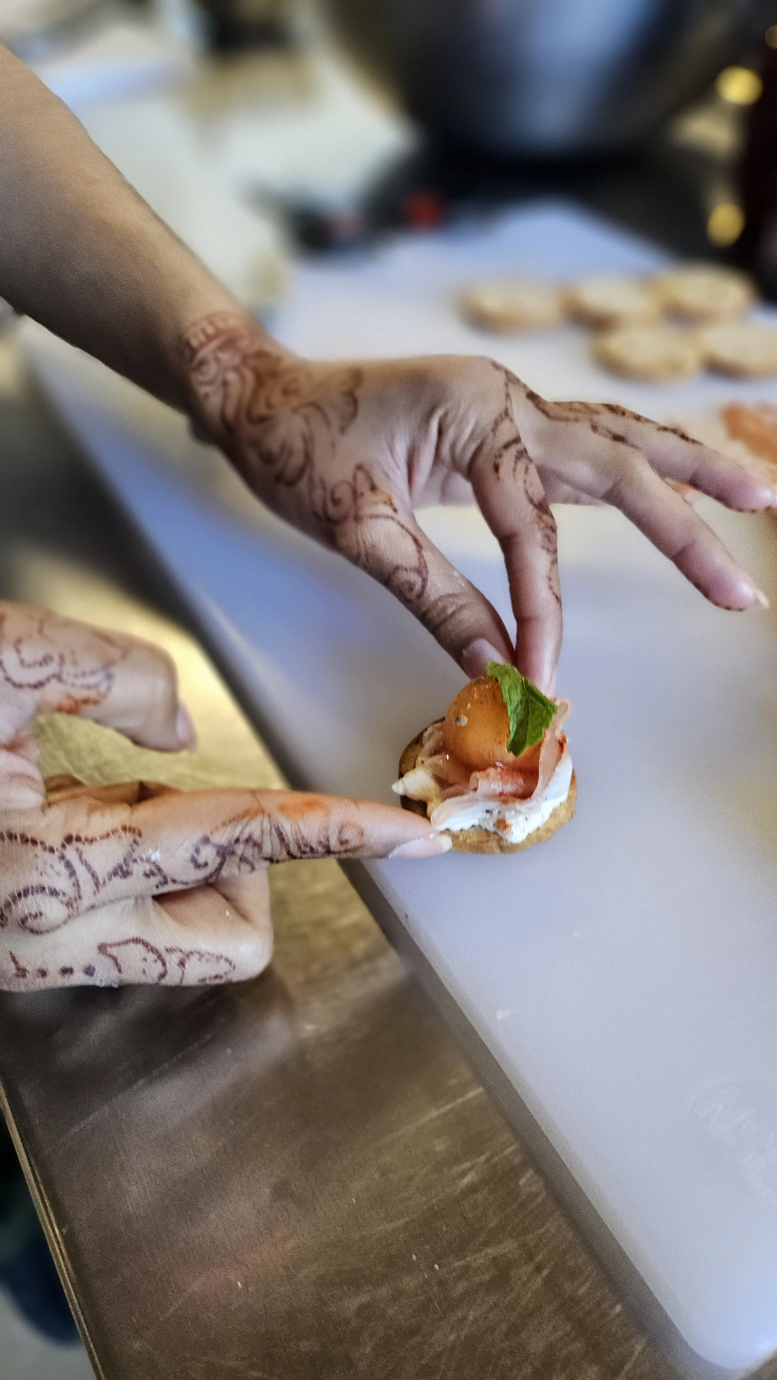 Person adding a garnish, possibly a cherry tomato and a leaf, onto a small canapé with cream cheese on a cookie or cracker, with other canapés in the background.
