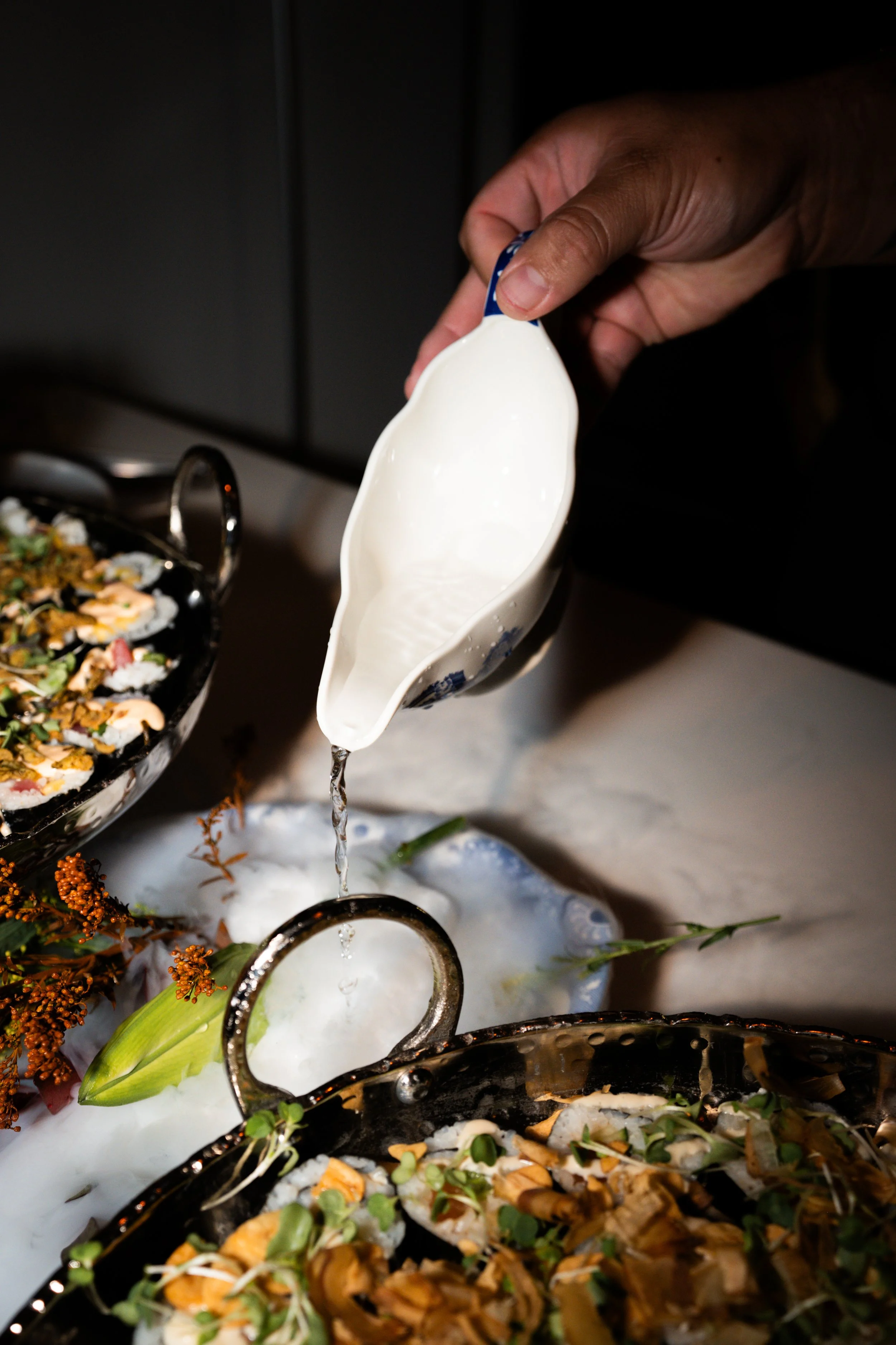 Someone pouring a white sauce or dressing into a serving dish at a food presentation.