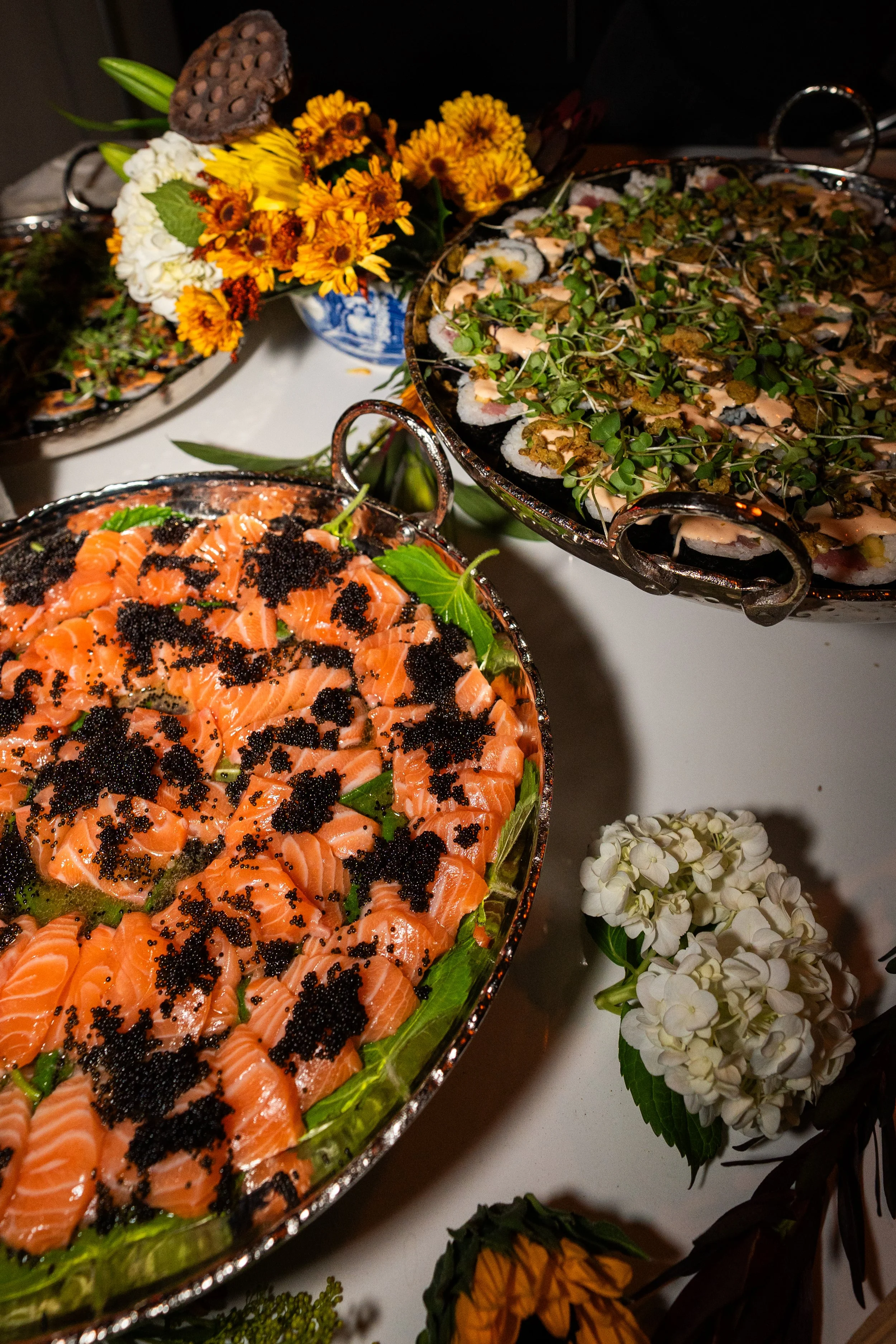 Plate of sliced salmon topped with black caviar, two silver trays of sushi rolls with drizzled sauce, and a bouquet of yellow, orange, white, and green flowers.