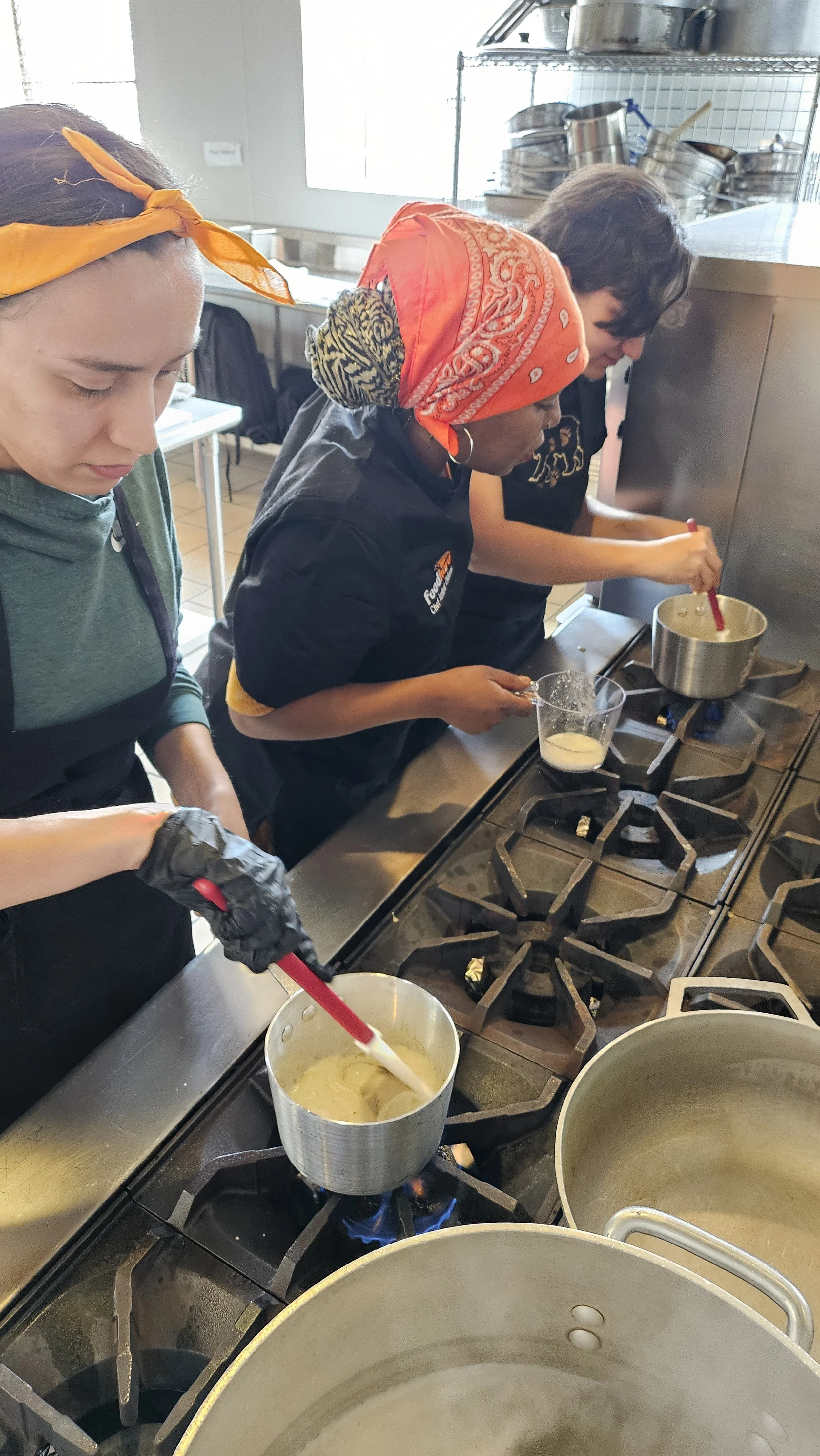 Three women cooking and stirring food over a stovetop in a professional kitchen. They are focused on their tasks, and one of them is wearing gloves while stirring a pot, another is holding a measuring cup, and the third is stirring a pot with a red s