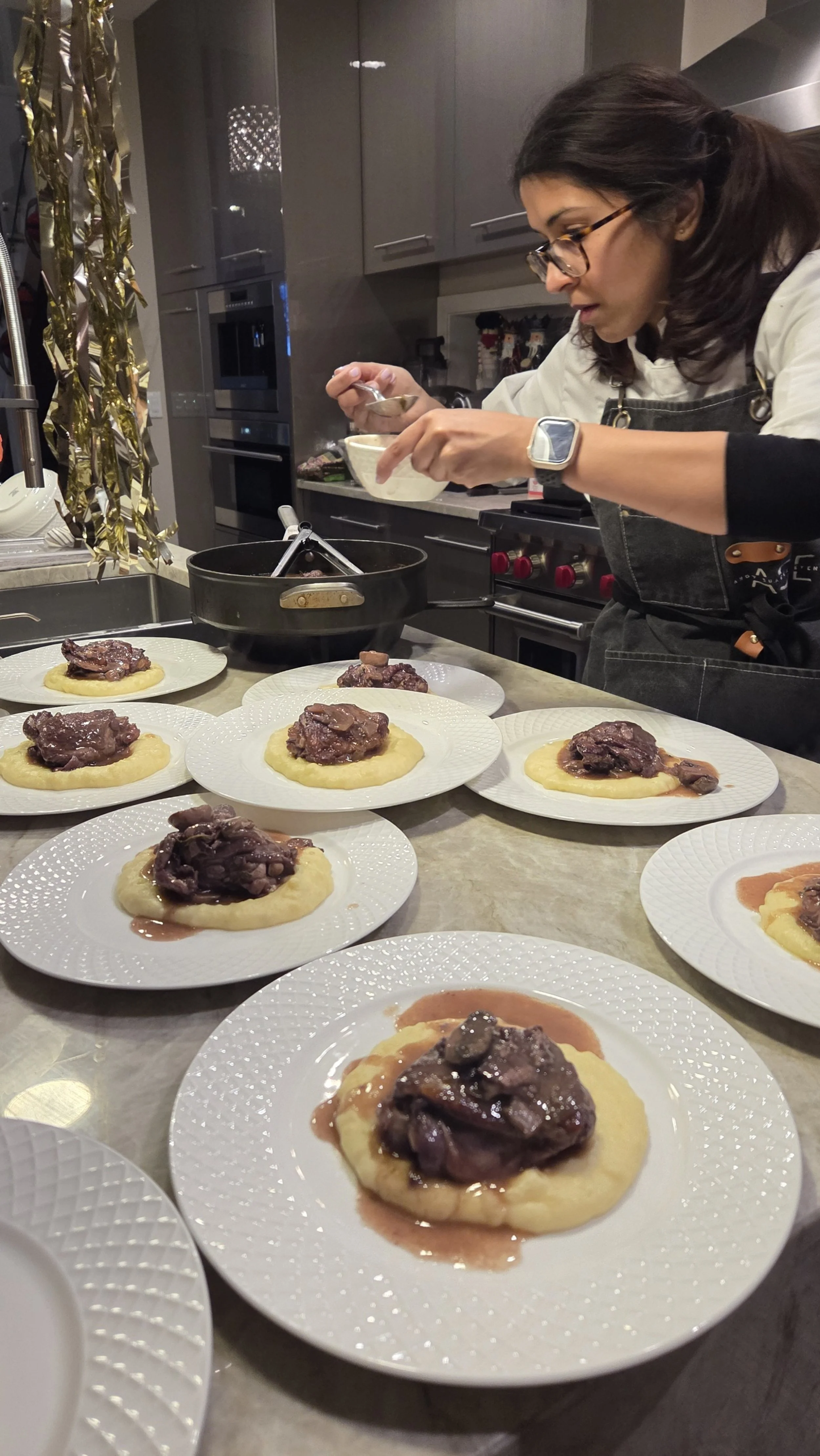 A woman preparing multiple plates of mashed potatoes topped with braised meat in a kitchen.