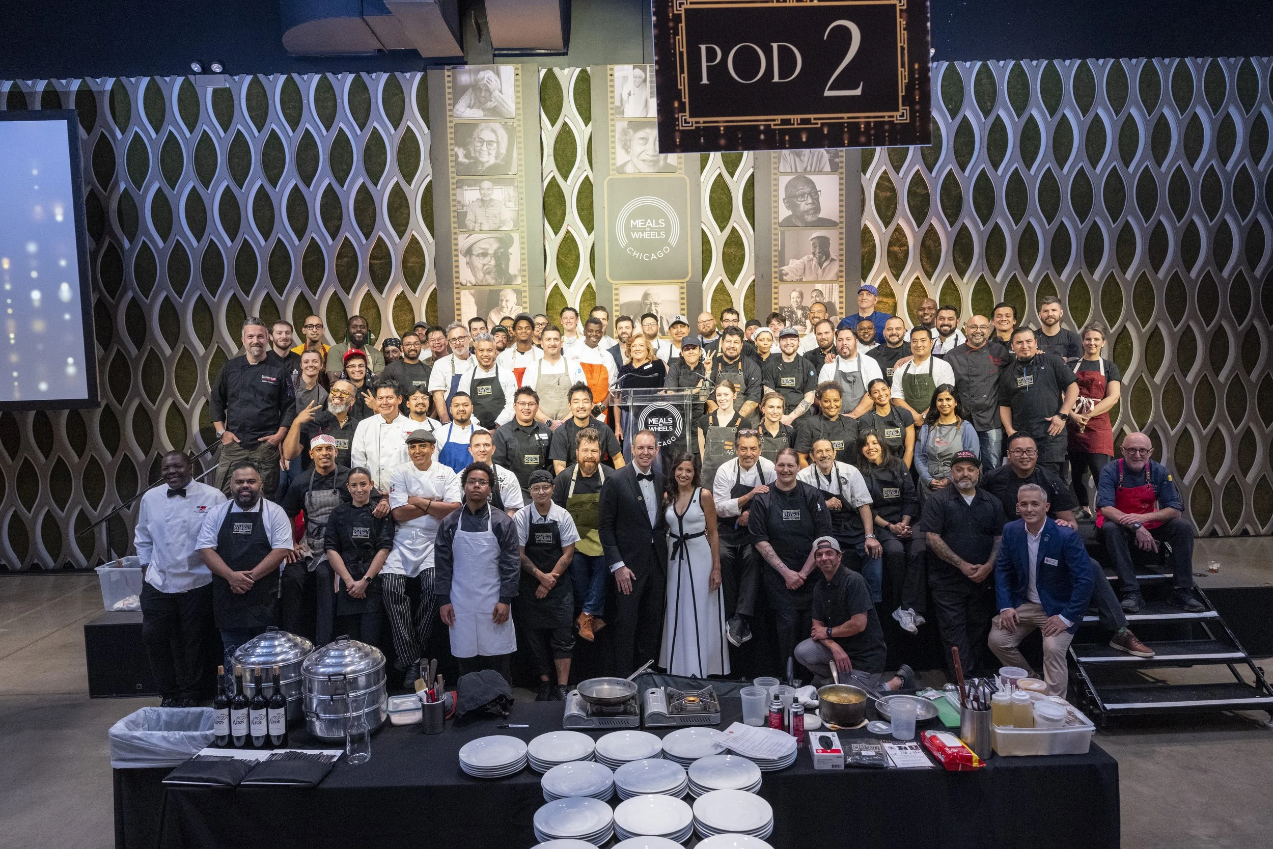 A large group of chefs, volunteers, and organizers posing on a stage at a cooking event, with a sign reading 'Pod 2' hanging above them. The background displays a decorative wall with photos and a logo that says 'Meals Wheels Chicago'. In front of th