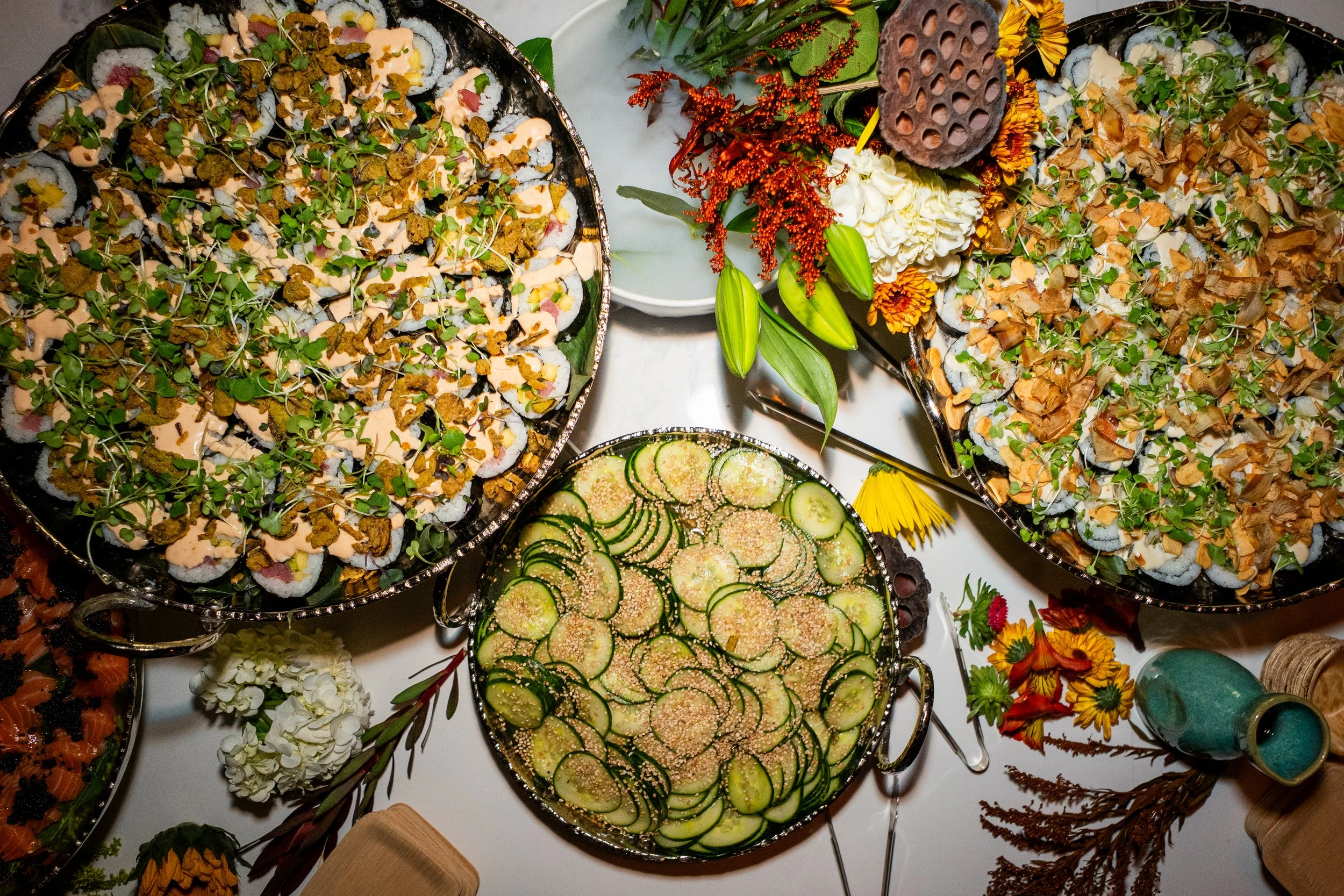 A table of assorted sushi rolls with various toppings, a cucumber salad garnished with sesame seeds, and decorative fall flowers and foliage.