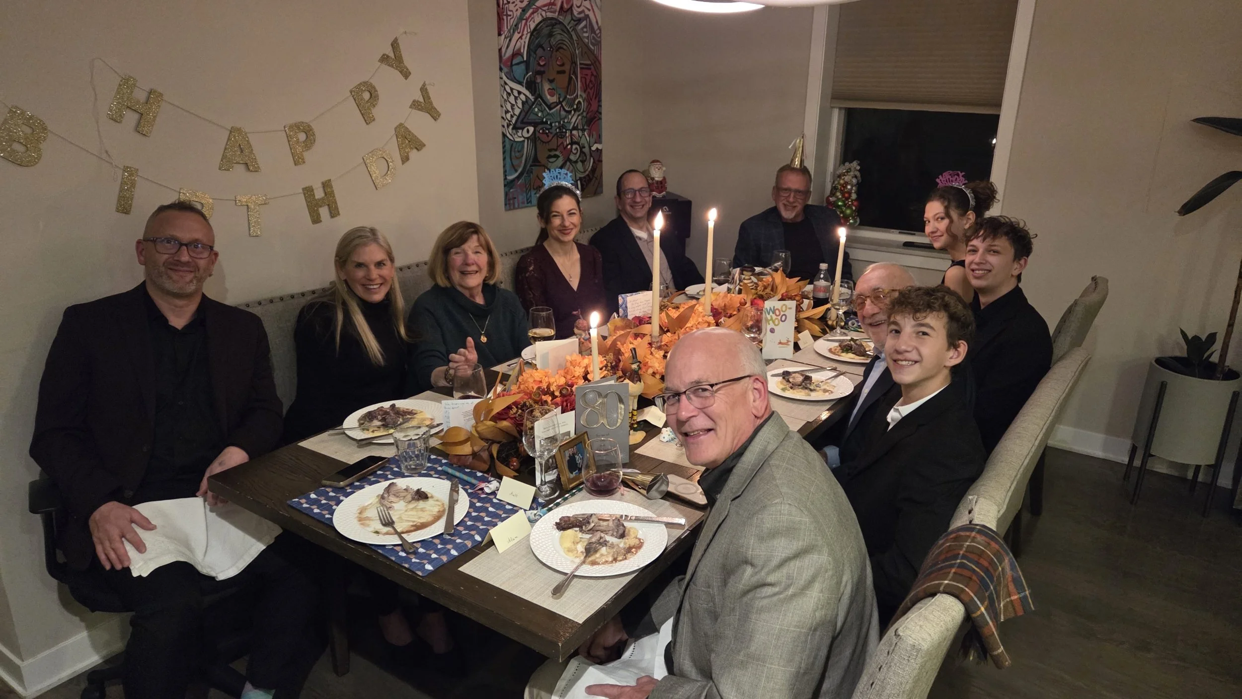 A group of eleven people sitting around a dinner table celebrating a birthday and a holiday, with a "Happy Birthday" banner on the wall, candles, and festive decorations.