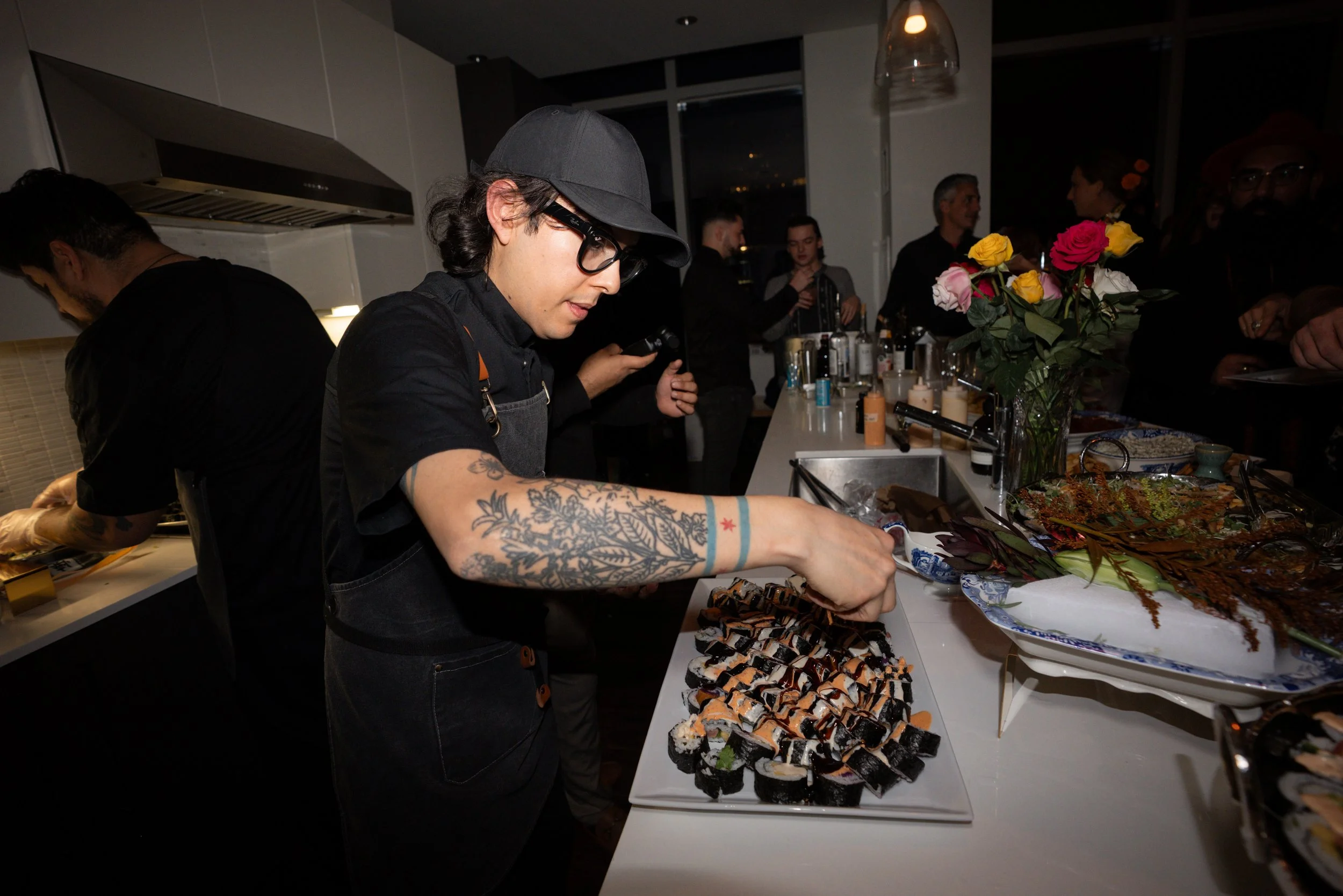A sushi chef with tattoos and glasses preparing sushi at a party or event, with guests in the background and a vase of flowers on the counter.