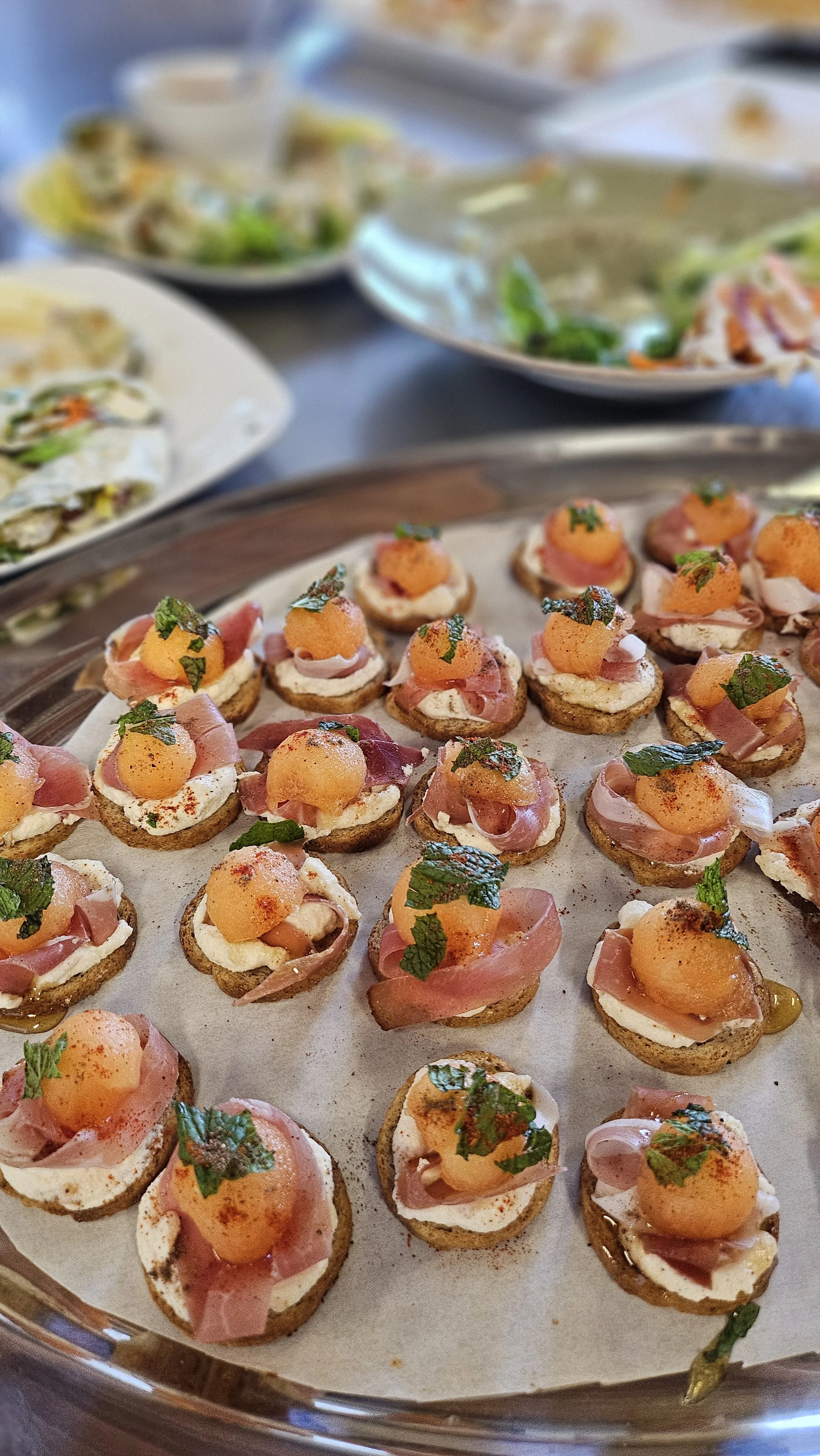 Tray of mini open-faced sandwiches with melted cheese, prosciutto, melon, and herbs, garnished with chopped fresh herbs.