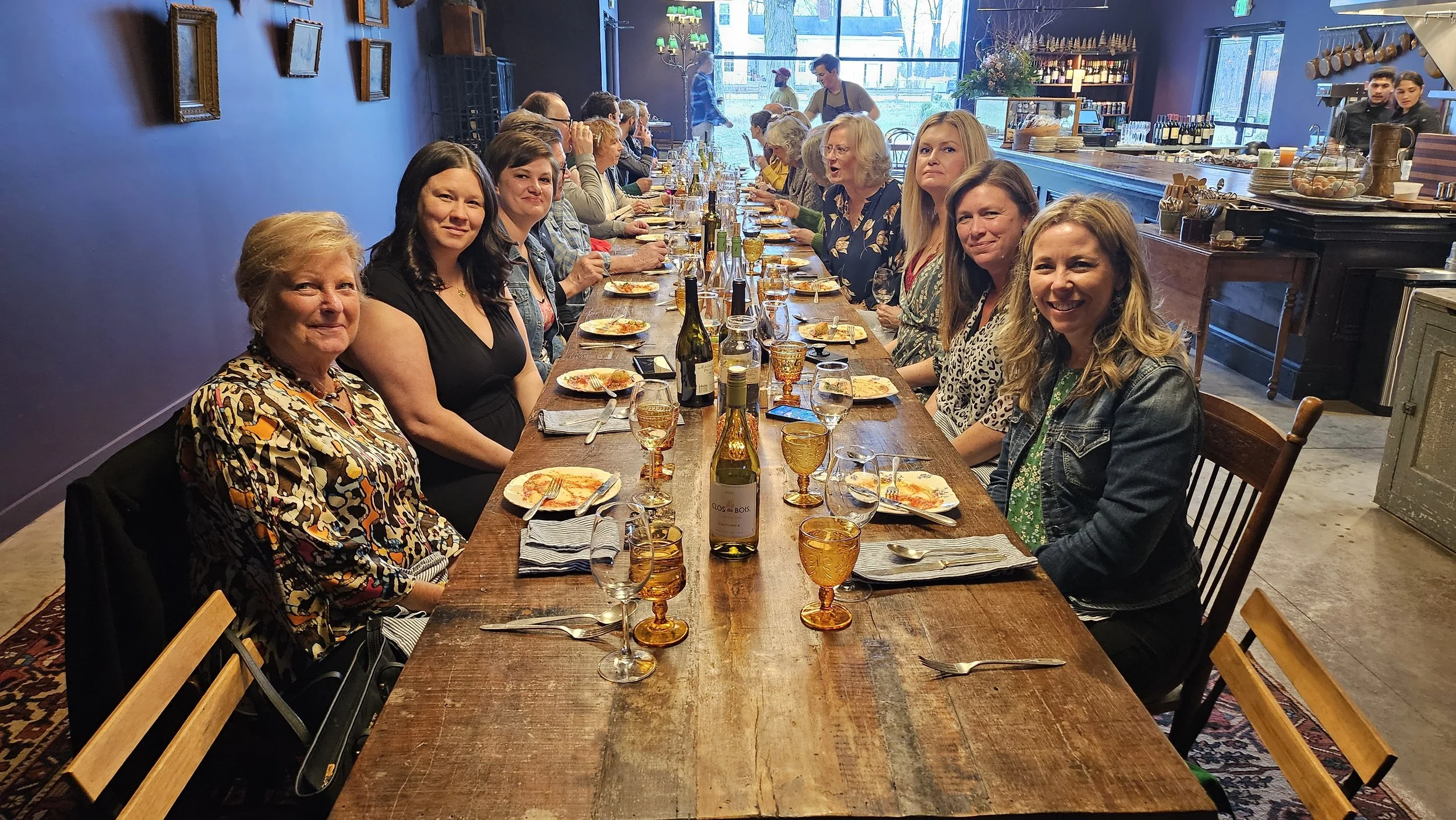 A large group of women sitting at a long dining table inside a restaurant, with plates, glasses, and bottles on the table, smiling and looking at the camera.