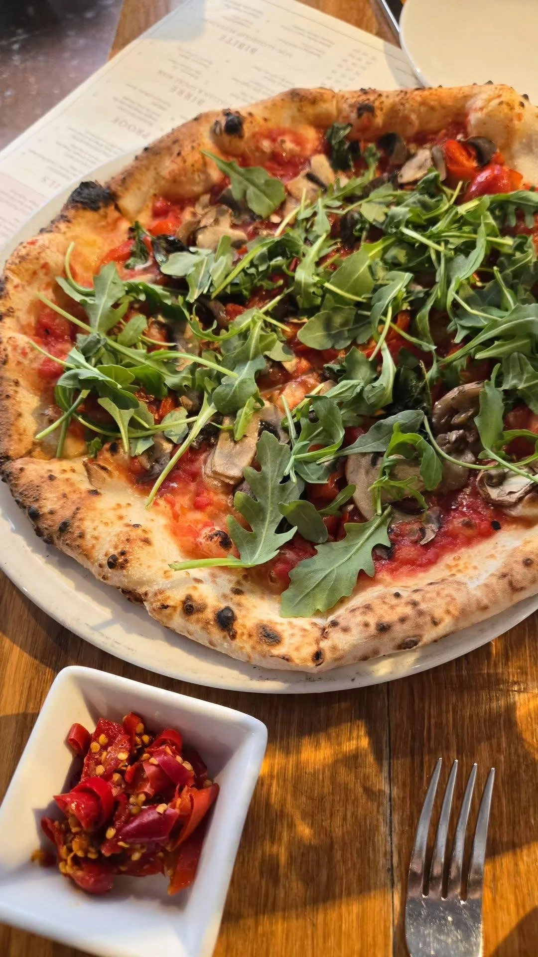 A pizza topped with arugula, mushrooms, tomatoes, and cheese on a wooden table, with a side of pickled vegetables in a small white dish and a fork next to it.