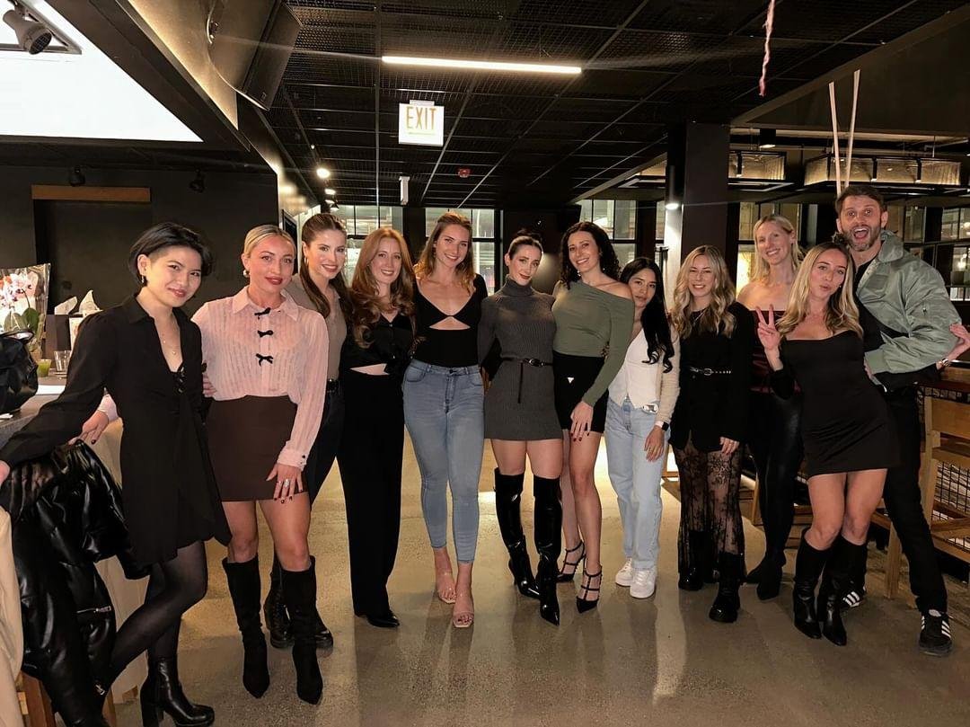 A group of fifteen people, mostly women, posing inside a dimly lit restaurant or bar. They are smiling, standing close together, with some making peace signs. The group appears happy and festive.