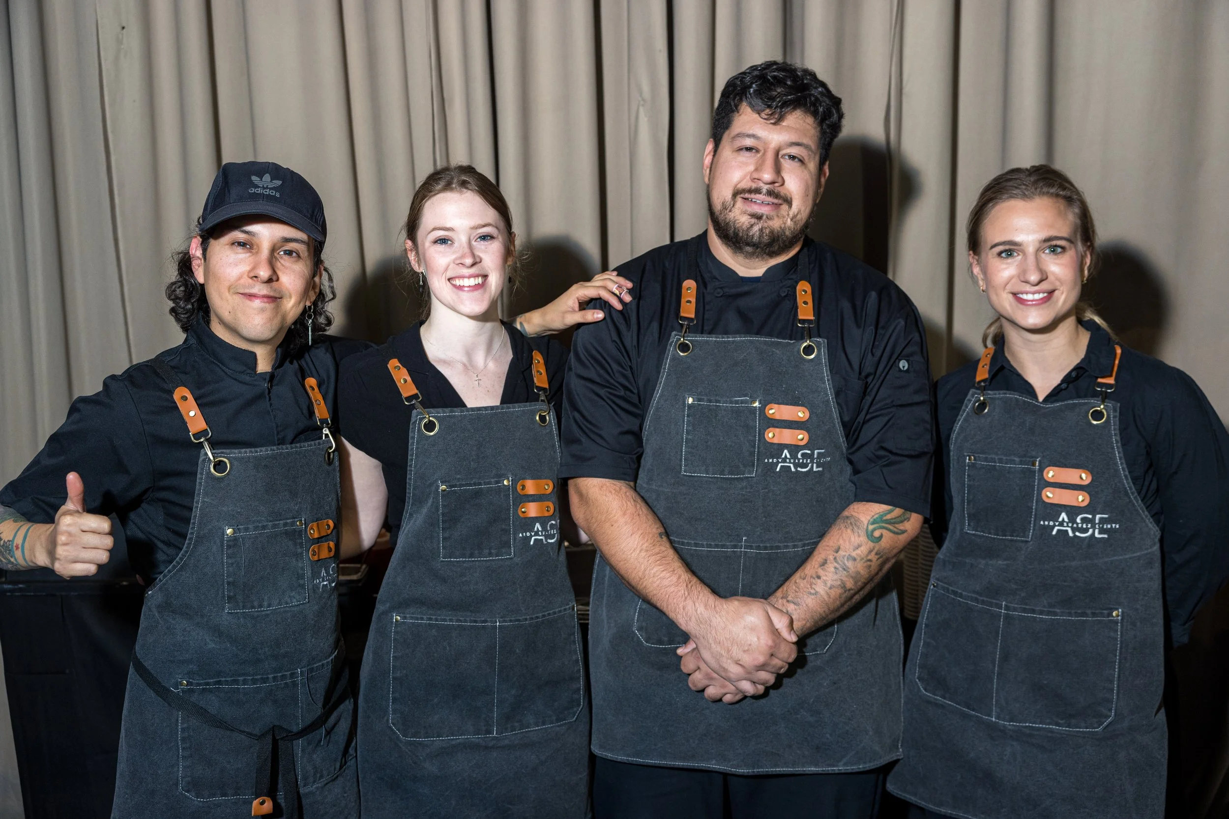 Group of four people wearing black chef uniforms and denim aprons, standing in front of beige curtains, smiling at the camera.