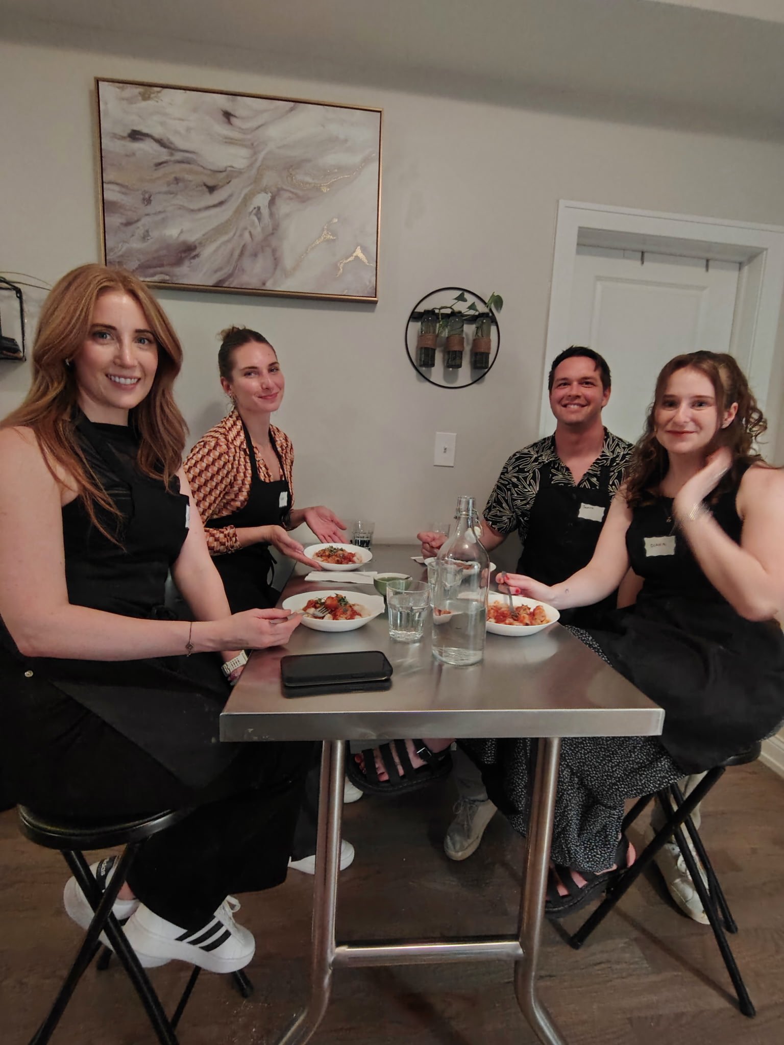 Four people sitting around a table with bowls of food, smiling, in a room with a wall art and decorative wall hanging.