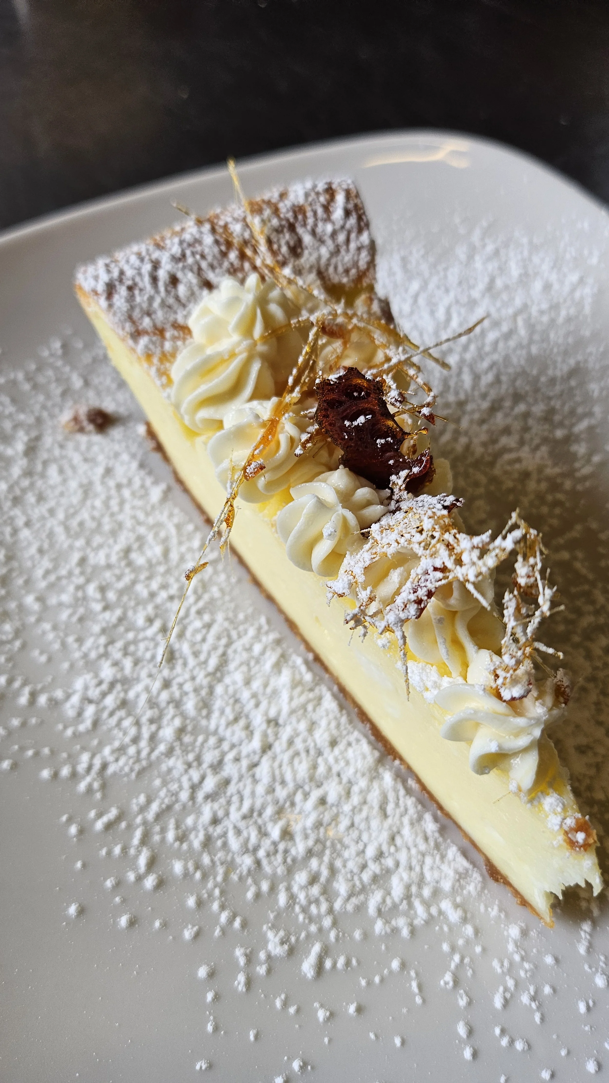 A slice of cheesecake garnished with whipped cream, caramelized sugar, and powdered sugar on a white plate.