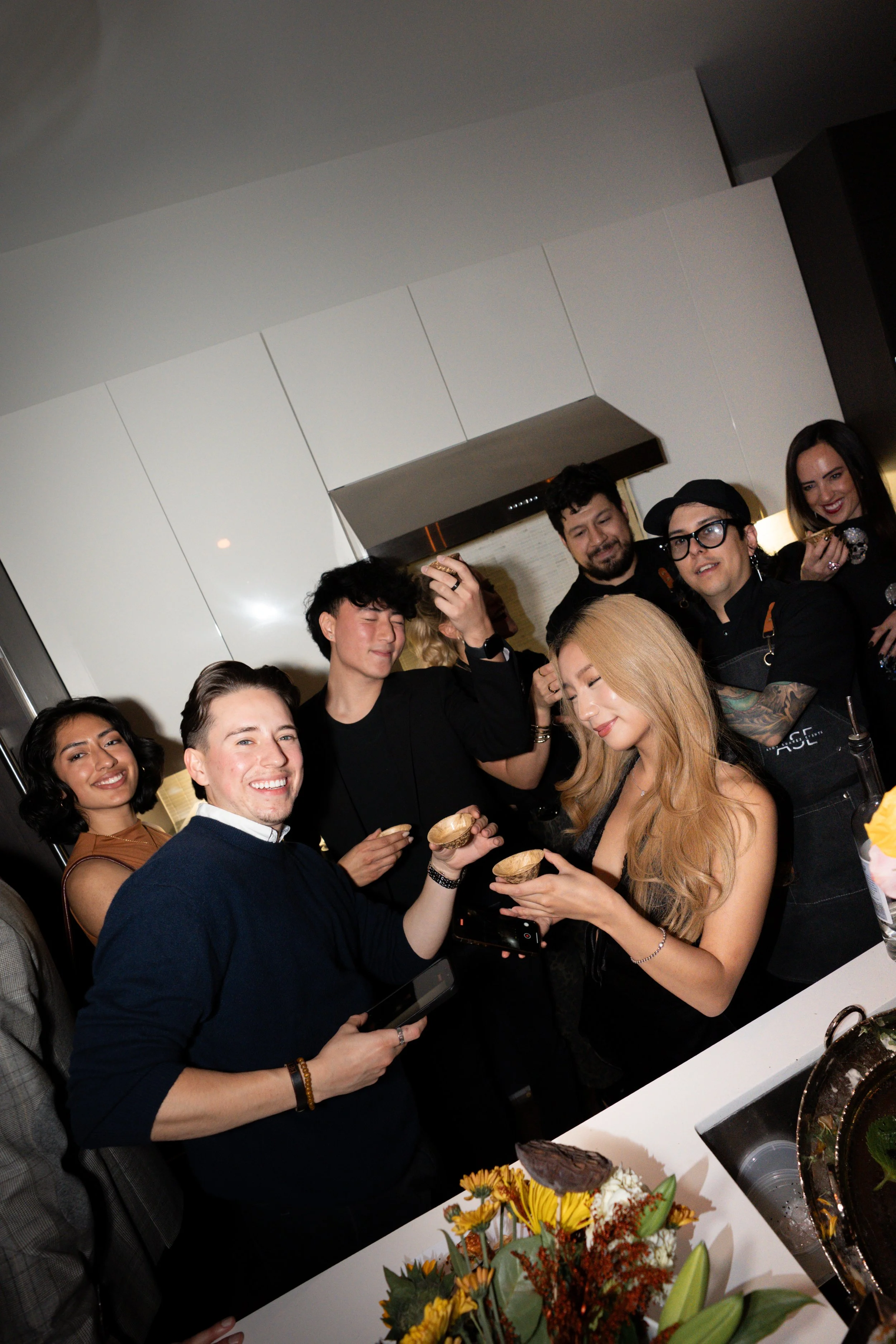 Group of friends celebrating in a kitchen, sharing desserts, smiling, with flowers and cake on the table.