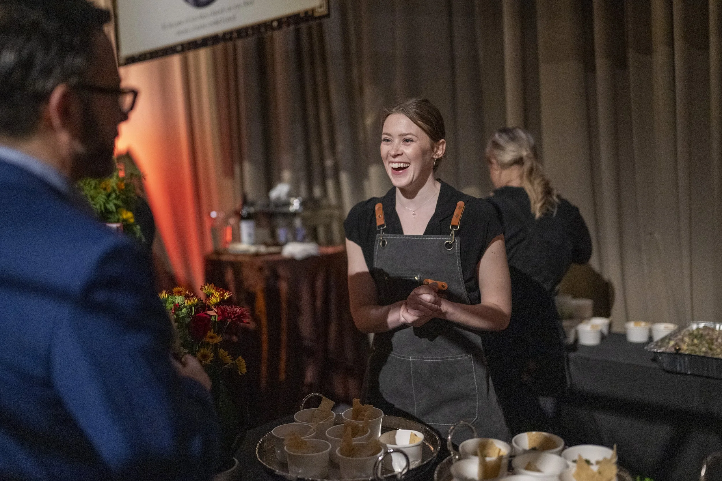 A woman with brown hair and a black apron is smiling and talking to a man holding a bouquet of flowers at a social event or party. There are small cups and snacks on the table in front of her, and a woman with blonde hair is in the background.