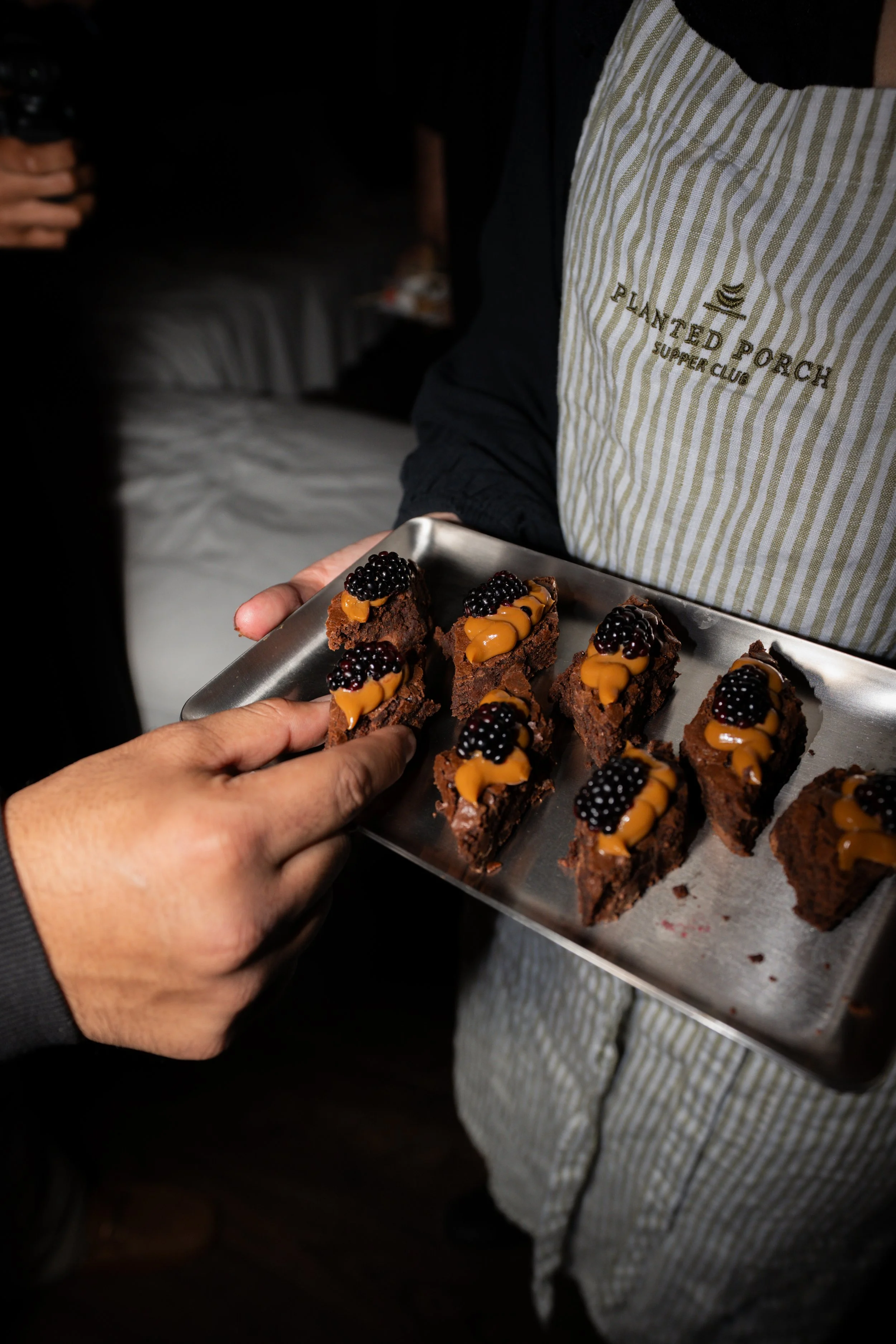 Person holding a tray of small chocolate brownies topped with blackberries and caramel drizzle, with another person in the background wearing a striped apron.