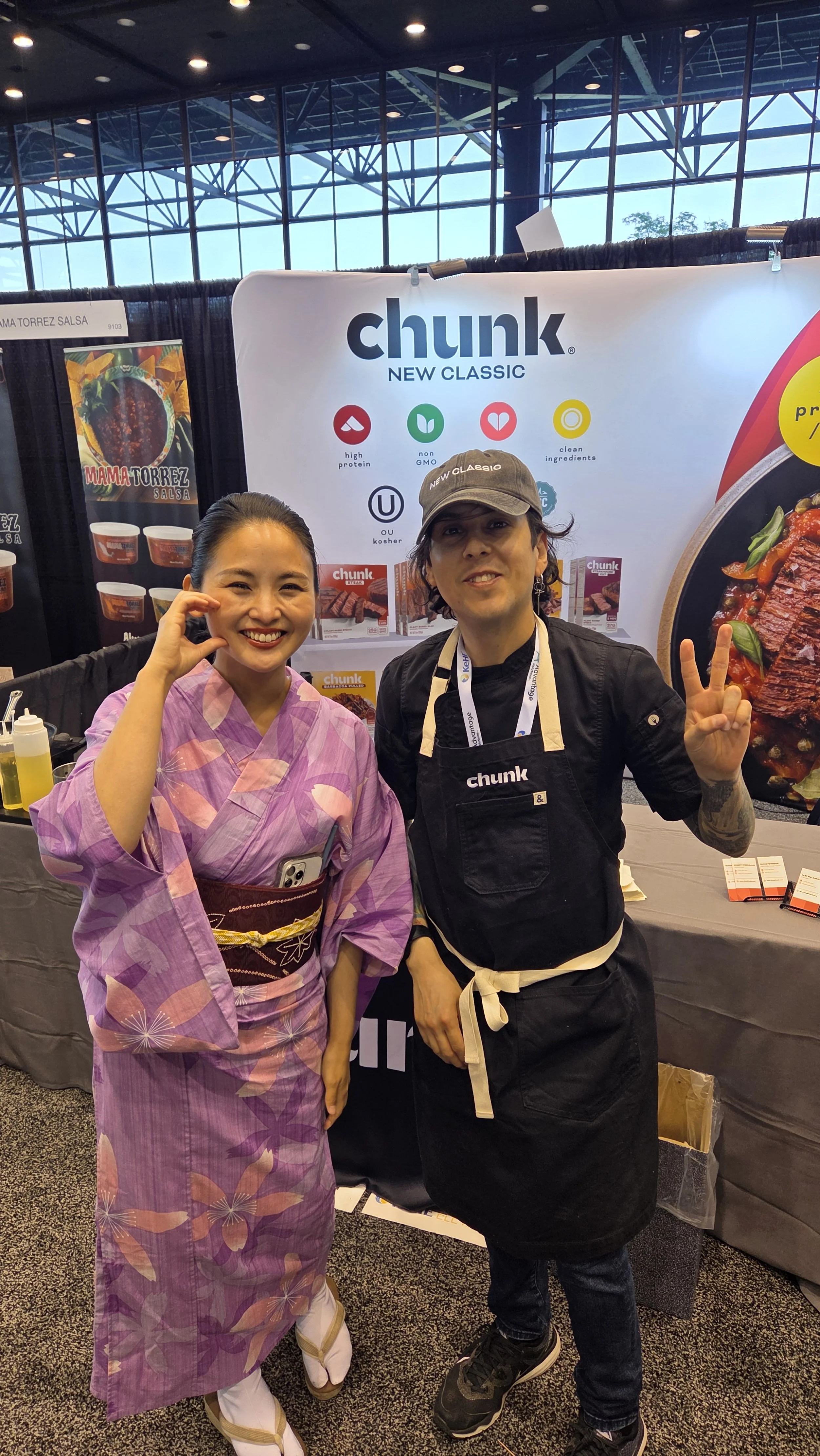 A woman in a pink kimono and a man in a black apron and cap posing and smiling at a booth at an event.