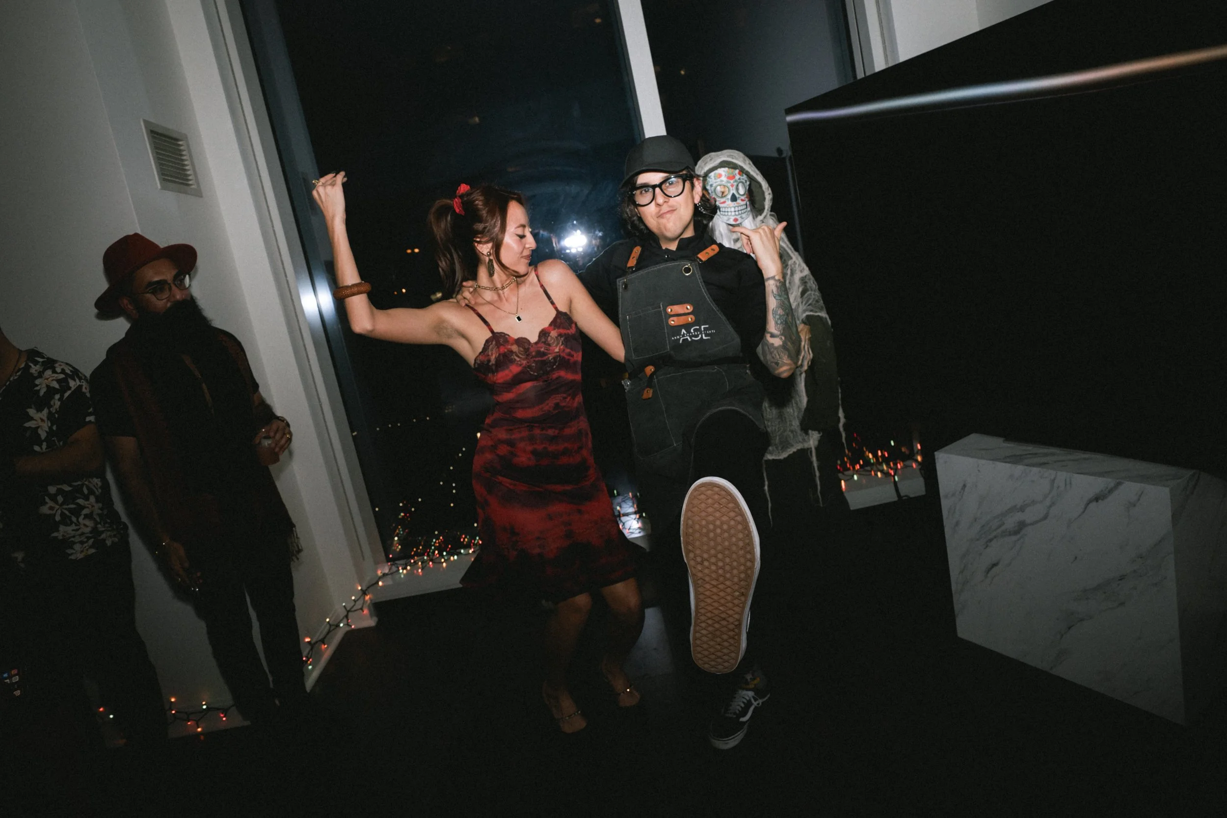 People dancing at a party near a large window at night, with Christmas lights visible outside.