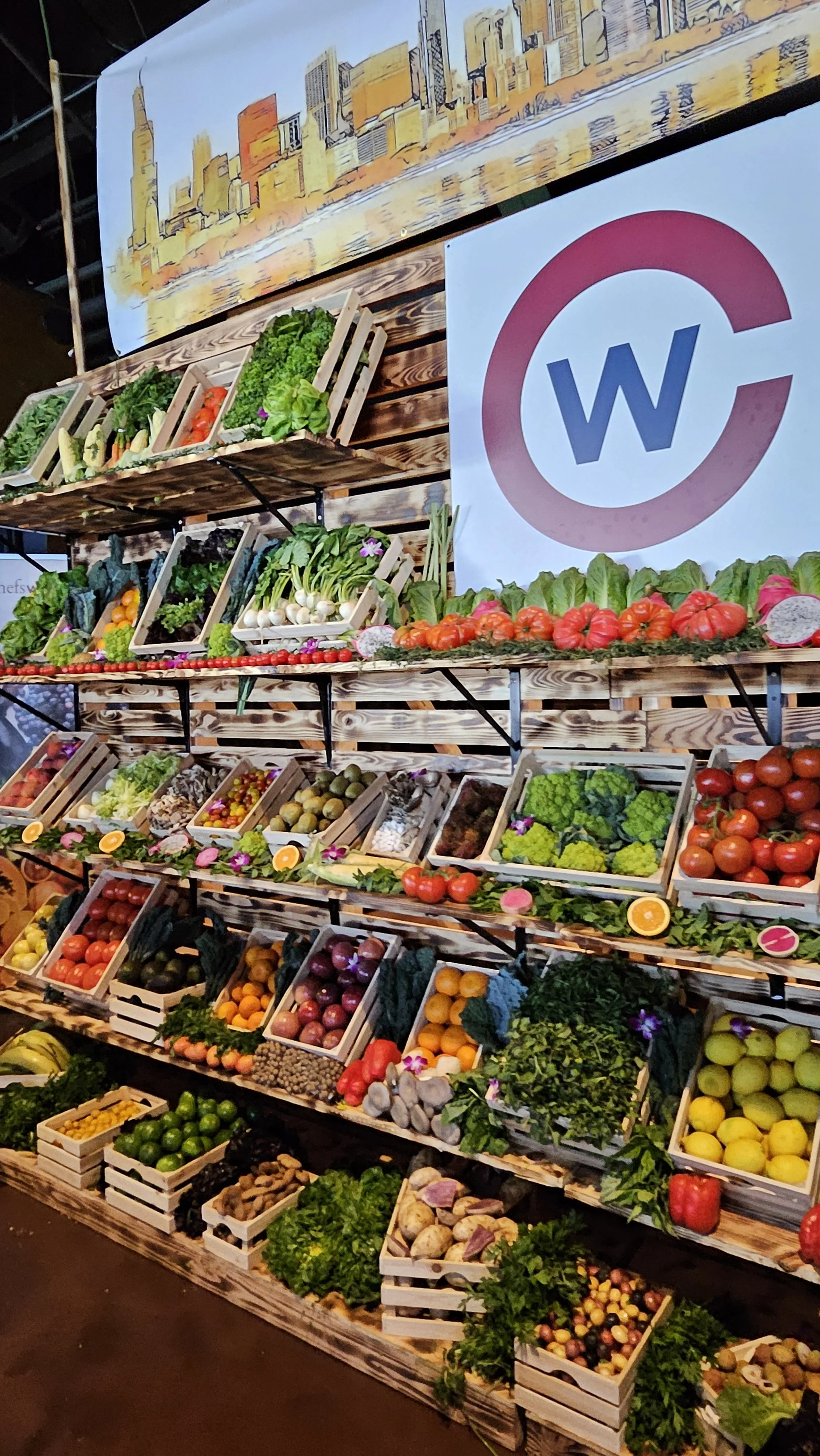 Display of fresh vegetables and fruits arranged on wooden shelves at a market stall with cityscape artwork and a logo with a red circle and blue 'W'.