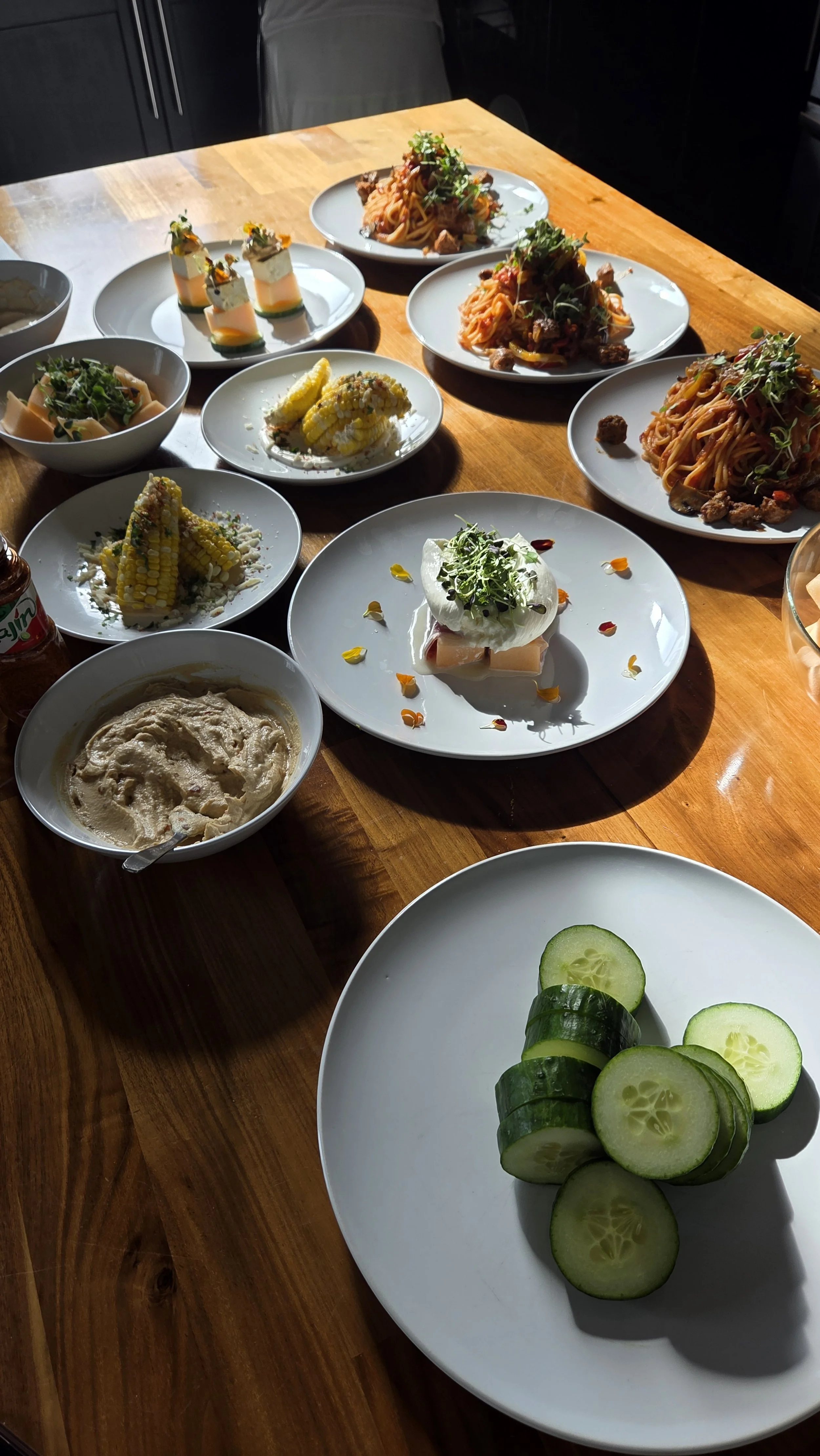 Assorted plates of food on a wooden table, including pasta dishes, corn on the cob, cucumber slices, and appetizers.