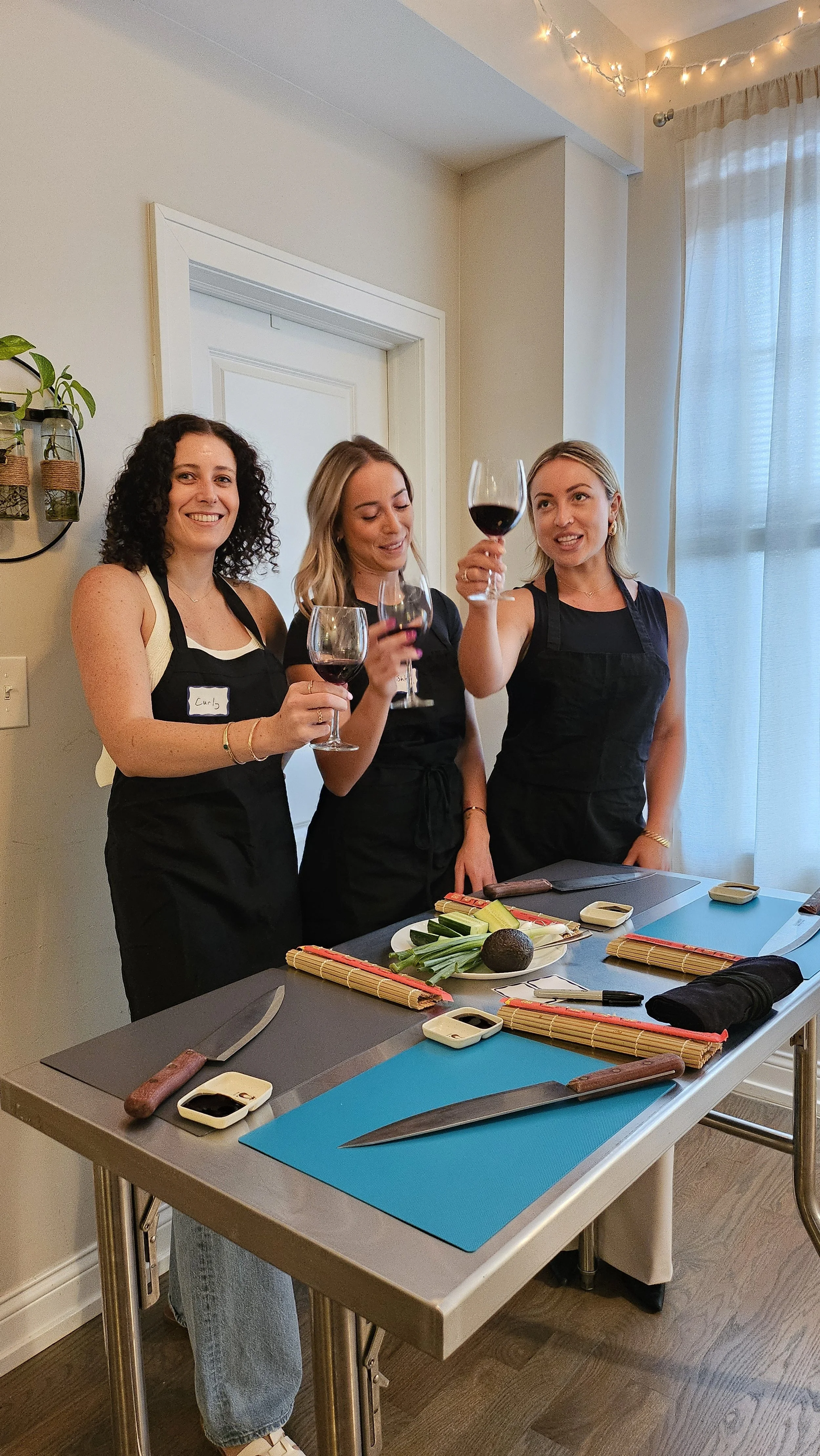 Three women wearing black aprons holding glasses of red wine in a kitchen or dining area with a table set for cooking or dining.