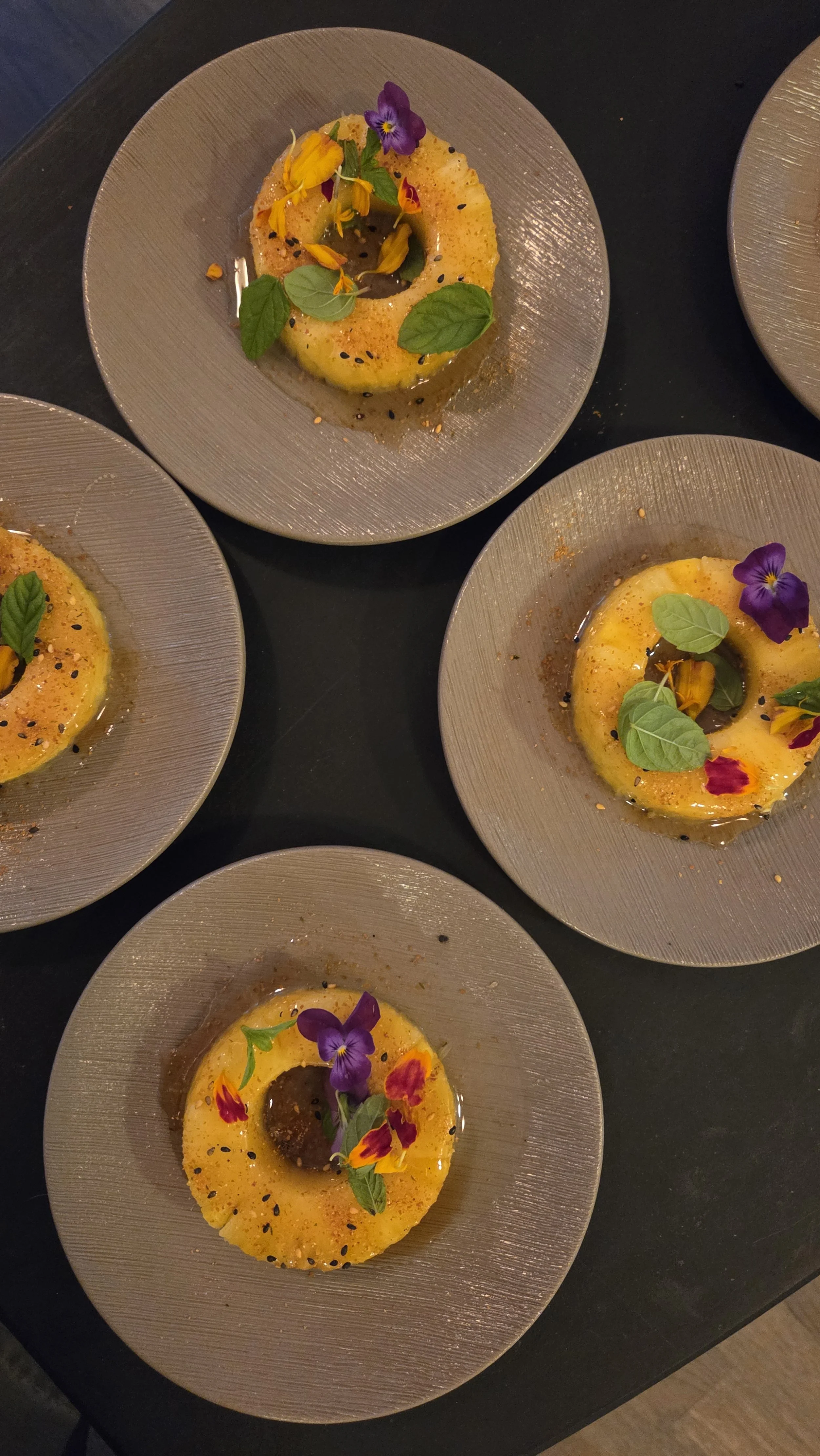 Four plates of yellow flan topped with edible flowers, green leaves, black sesame seeds, and orange crumbs on elegant gray dishes.