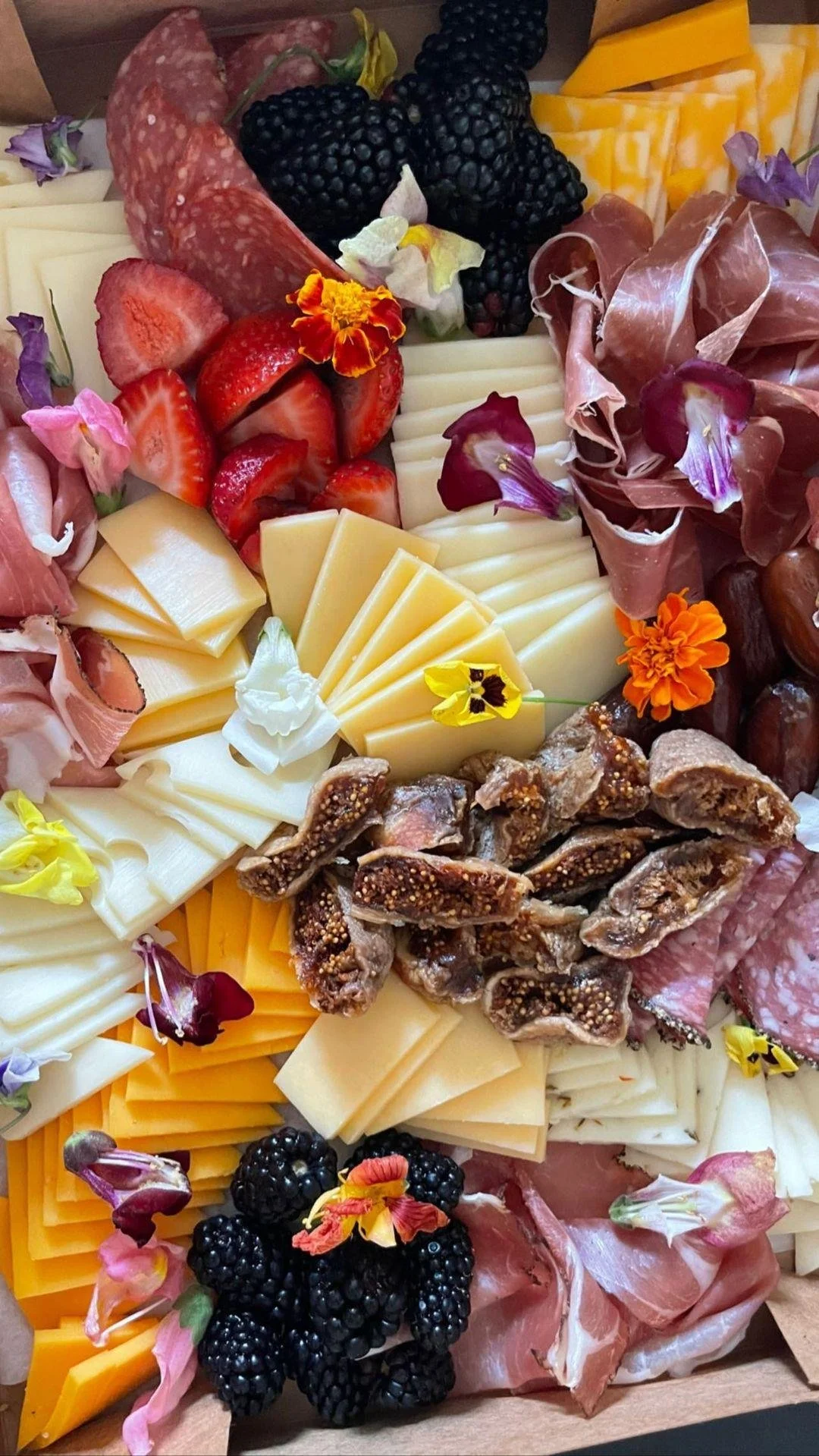 A cheese and charcuterie board with slices of cheese, cured meats, blackberries, strawberries, and garnished with edible flowers.