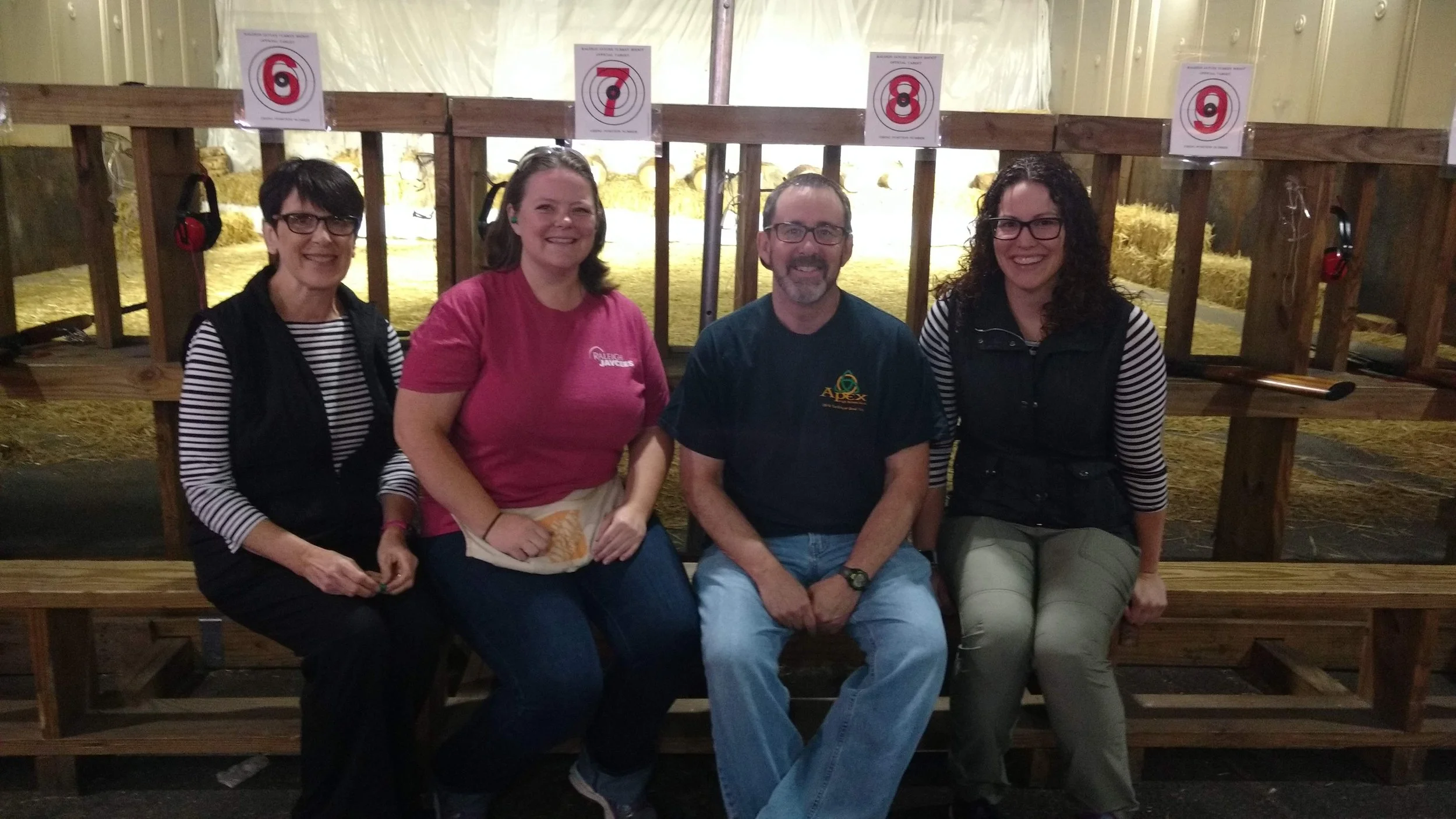 Four people sitting on a bench facing the camera. They are in front of an outdoor shooting range filled with hay.