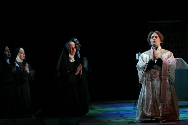 A scene from an opera with nuns and a priest kneeling and praying.