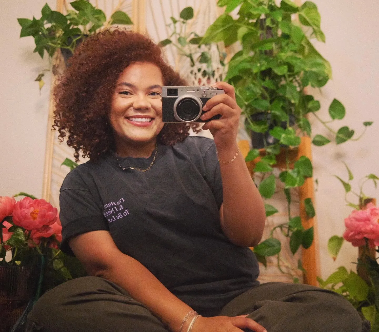 A smiling woman with curly hair taking a mirror selfie in a room filled with green plants and pink flowers.