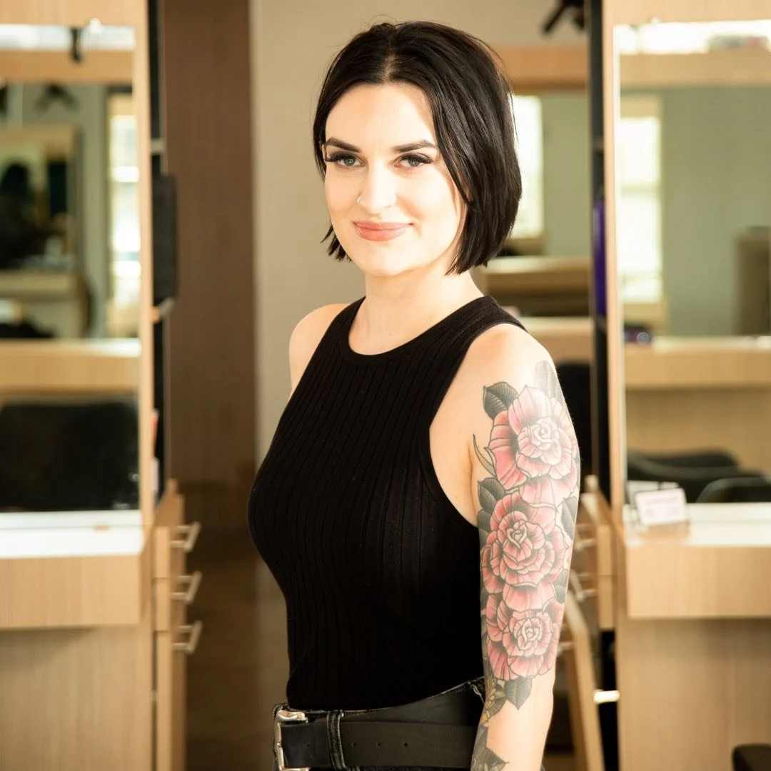 A woman with black hair and a black sleeveless top, smiling, showing a floral tattoo on her left arm, standing inside a modern salon or barbershop.