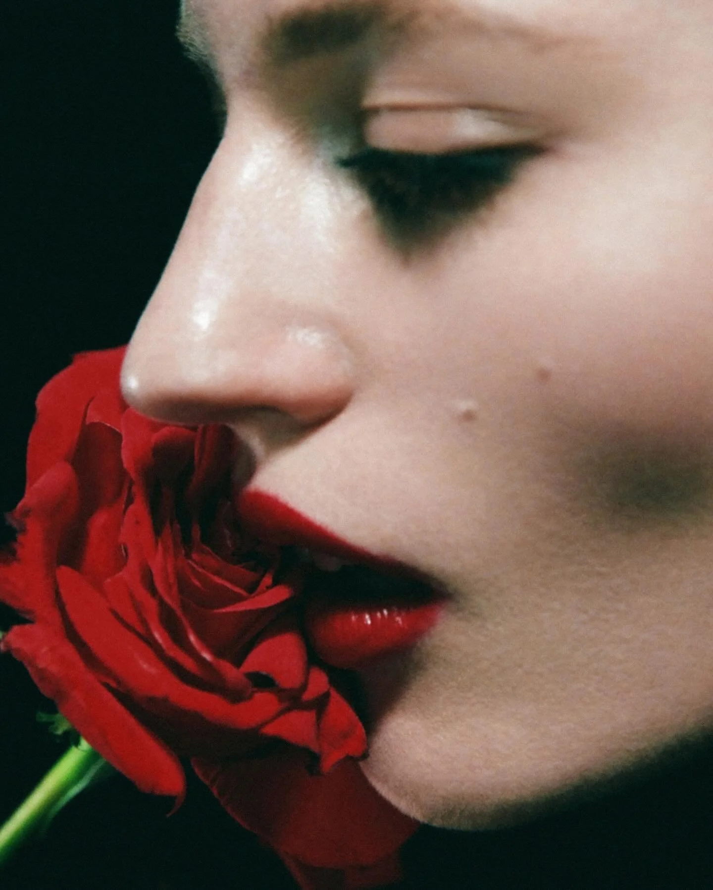 Close-up of a woman's face with red lipstick, holding a red rose near her lips with her eyes closed.