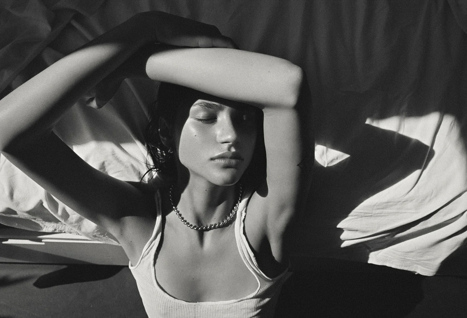 Black and white photo of a young woman lying on a bed with her arms raised above her head, wearing a tank top, a chain necklace, and closed eyes.