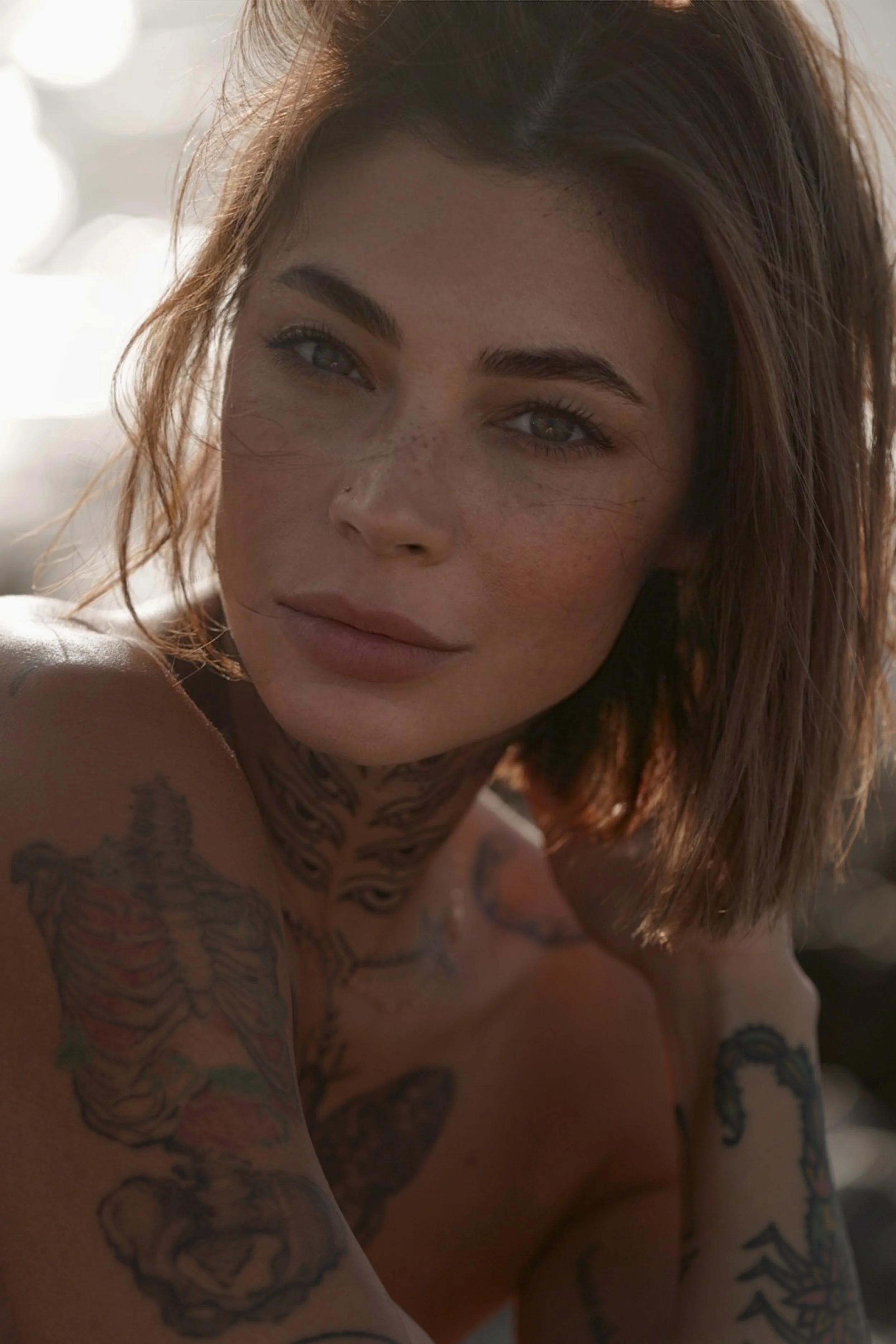 Close-up of a woman with short brown hair, freckles, and tattoos on her shoulders and chest, looking at the camera with a soft expression.