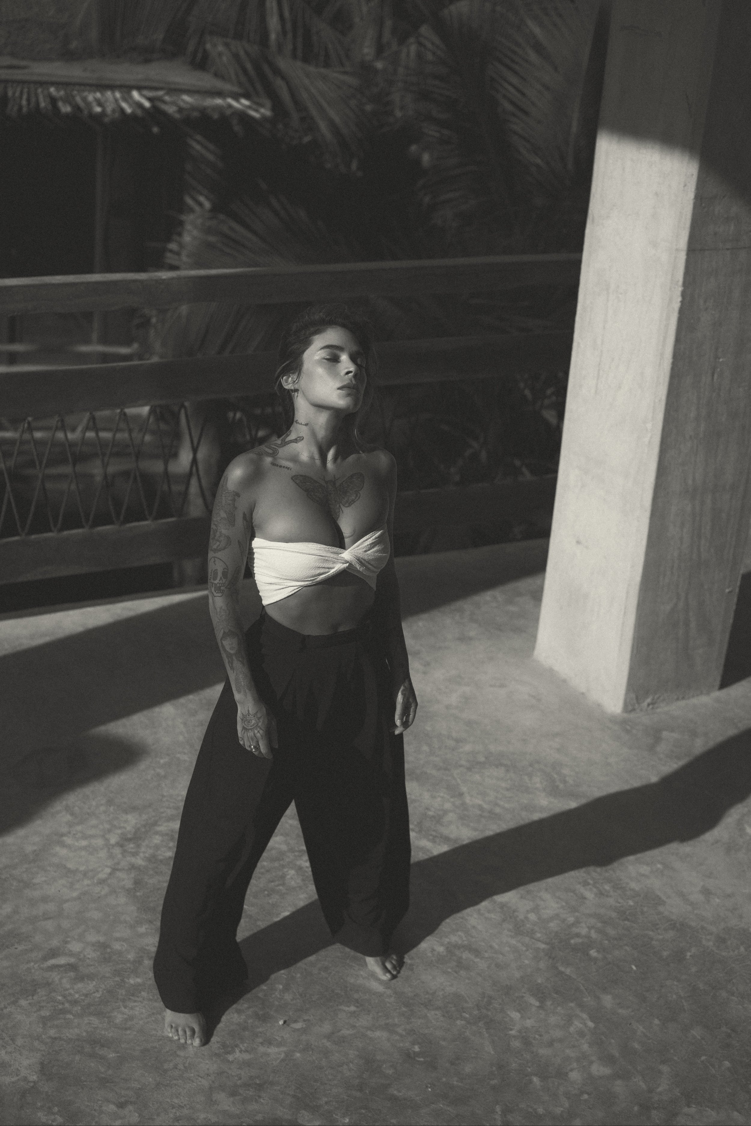 A tattooed woman with dark hair, wearing a strapless top and loose pants, standing outdoors in shadow with a textured concrete surface and architectural structure in the background.