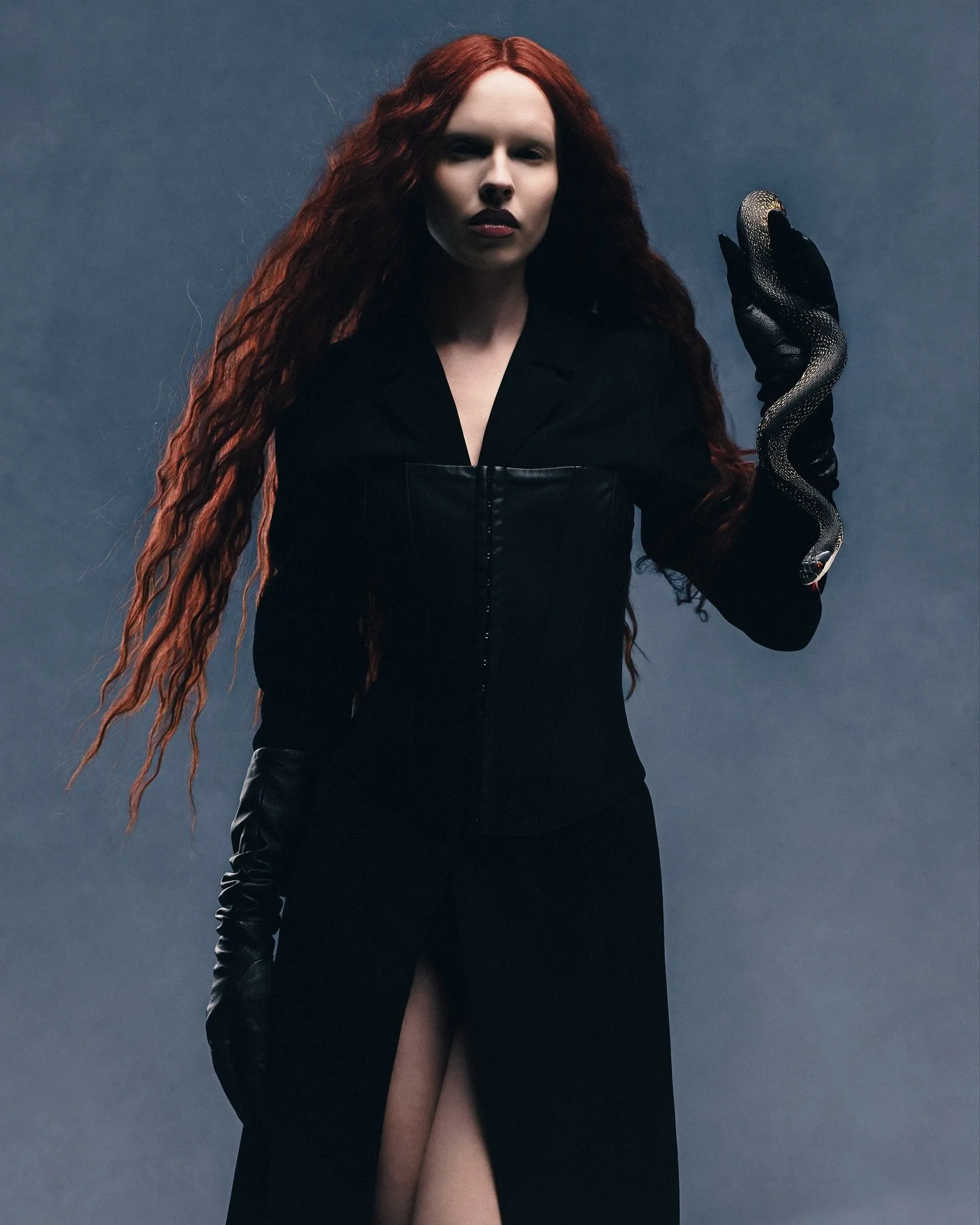 A woman with long, wavy red hair wearing a black dress with a slit, holding a black snake in her gloved hand, standing against a gray background.